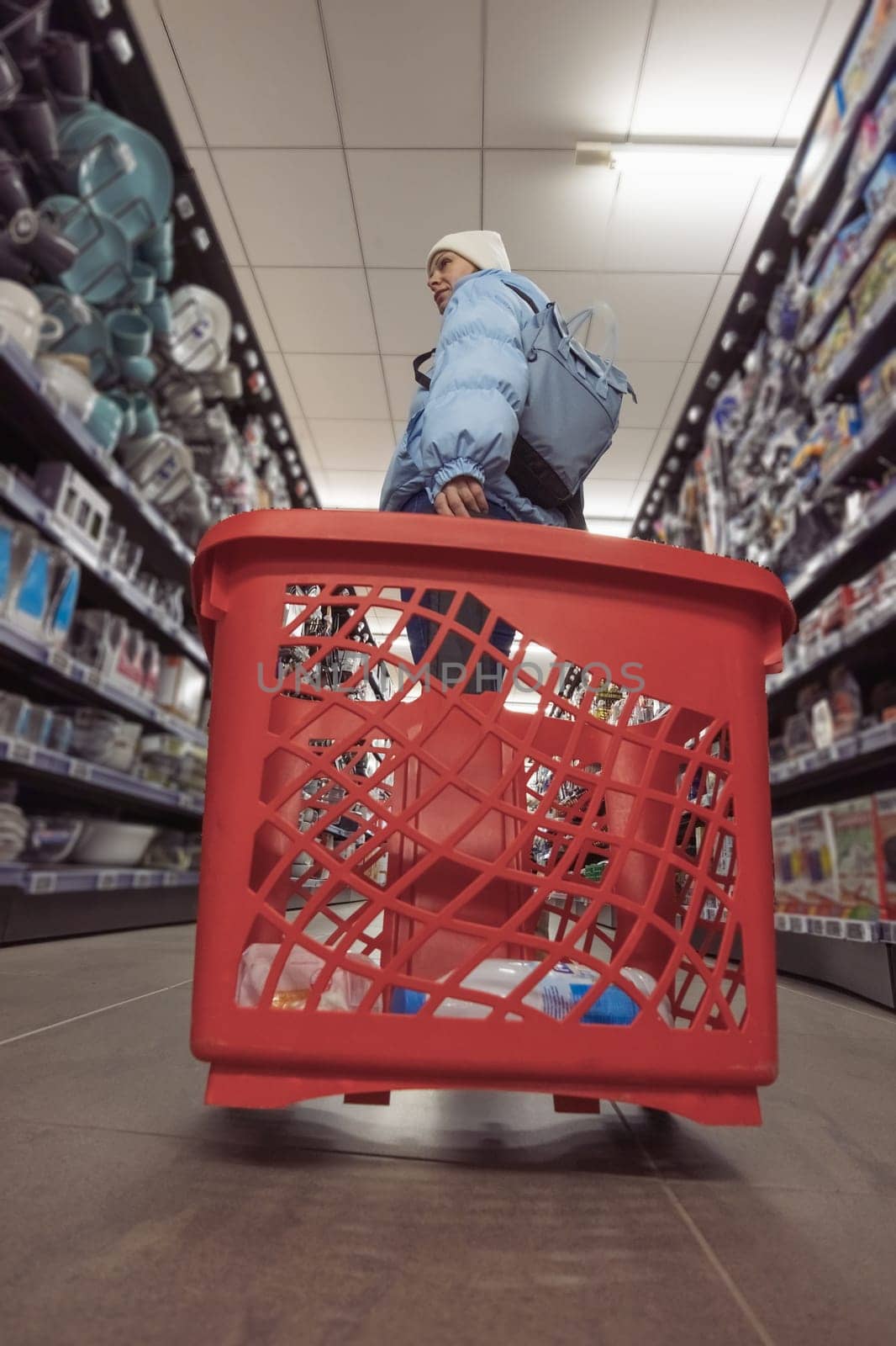 a girl of European appearance in a blue jacket and a white hat, standing in a store with a red cart, chooses dishes by PopOff