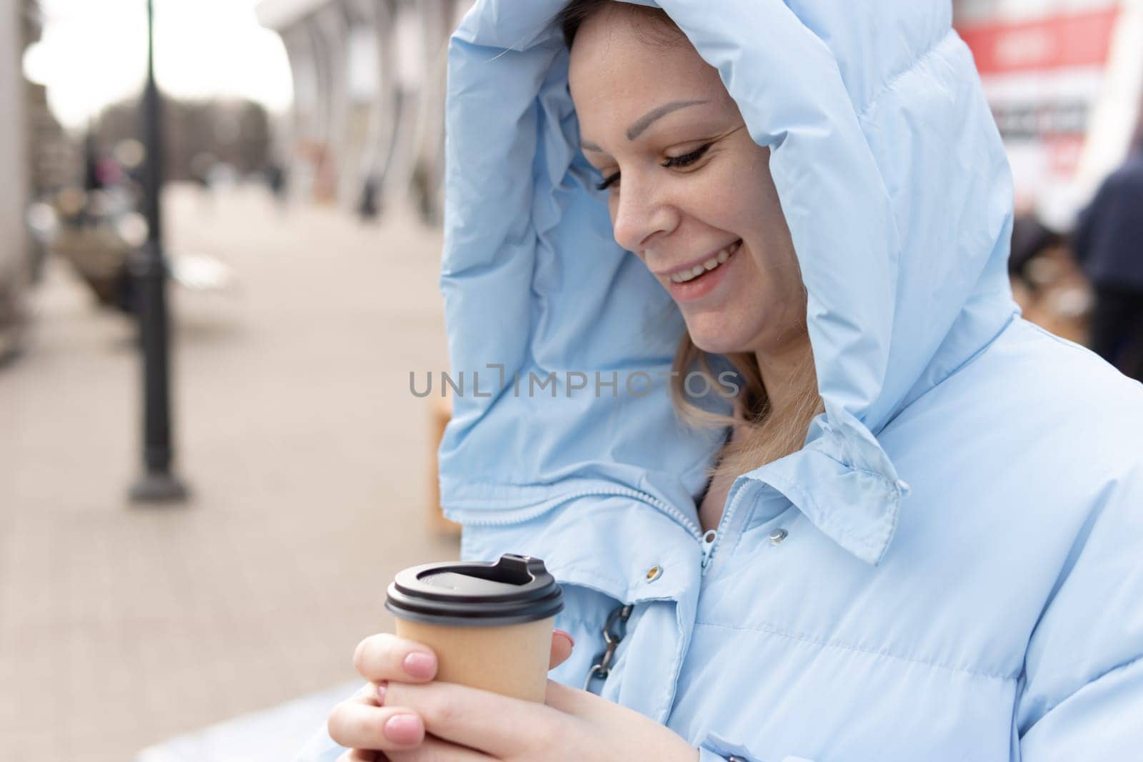 a girl of European appearance with blond hair, in a blue jacket, smiles and holds a paper cup of coffee in her hands. Stands on the street or in the park. Leisure concept. High quality photo