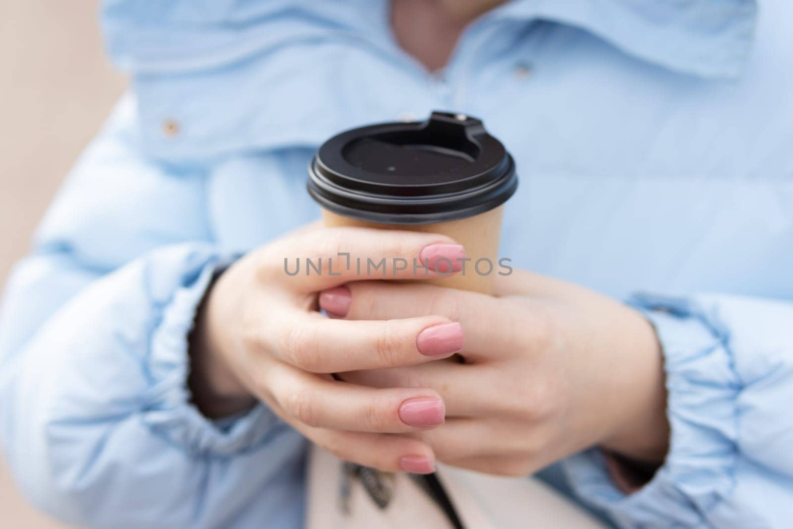 the girl is dressed in a blue jacket, with a white handbag holding a paper cup of coffee with her hands, close-up of a cup. High quality photo