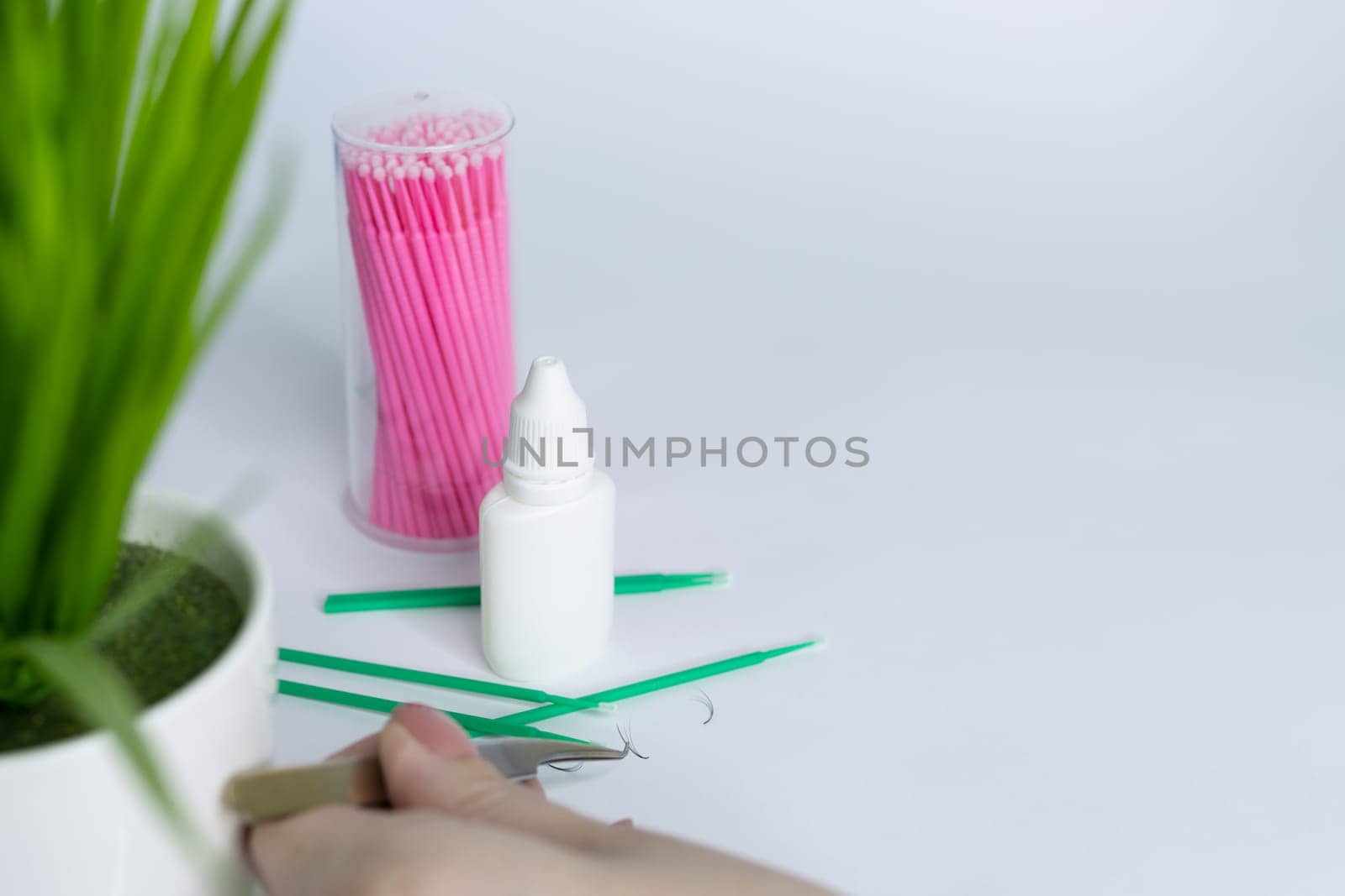 Woman's hand holds eyelash extension tweezers and takes artificial eyelashes. Materials for eyelash extensions lie side by side on a white background. On the right there is a place for an inscription