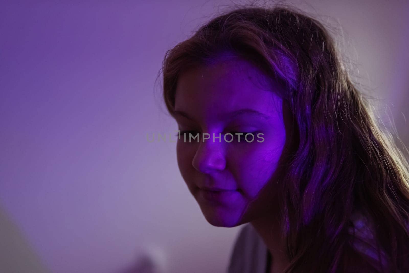 teenage girl of European appearance with blond hair, sits in a dark room with a colored light turned on, a purple light falls on the girl's face, there is a place for an inscription.High quality photo