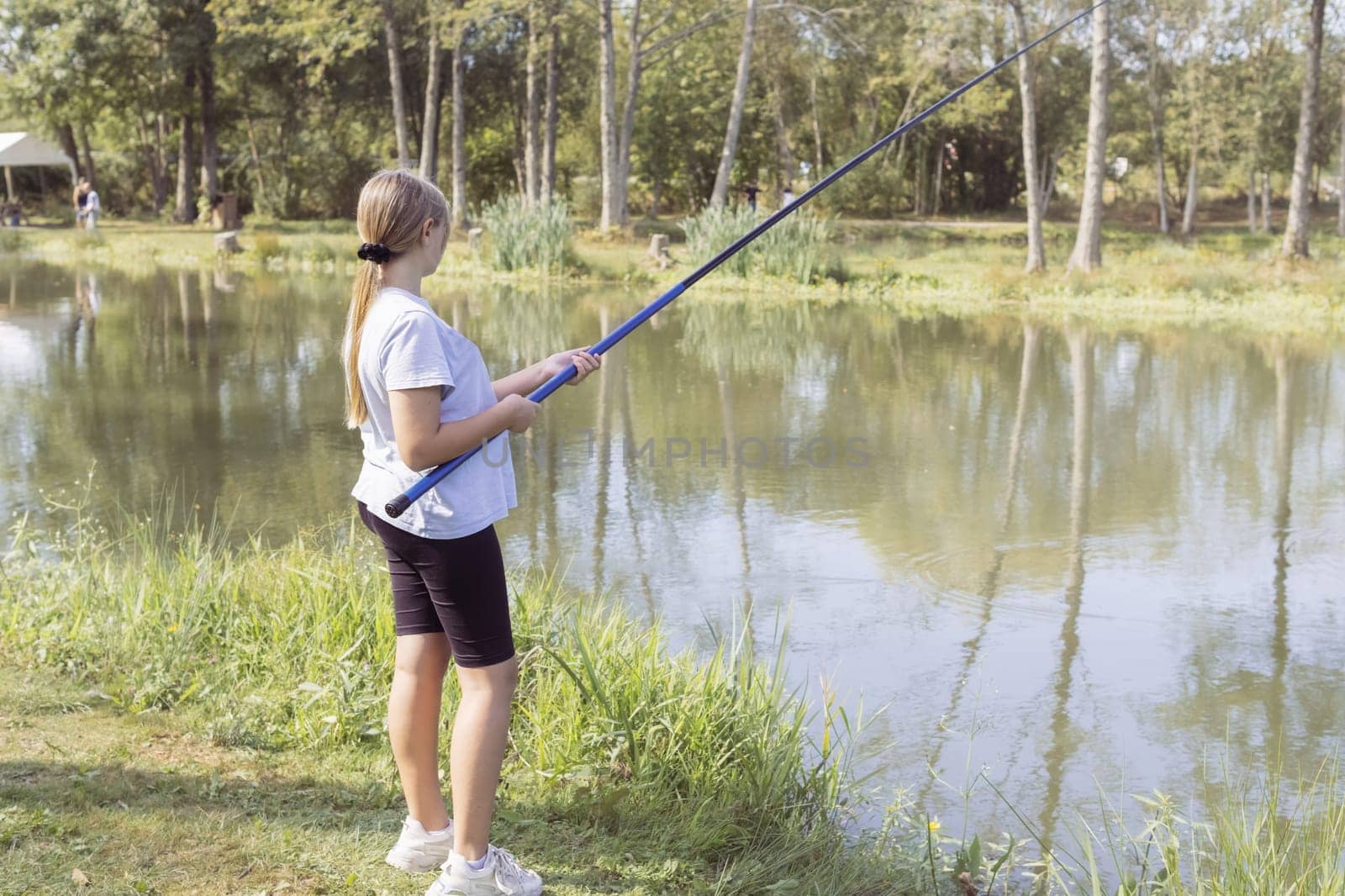 a teenager girl of European appearance with blond hair tied in a ponytail, stands fishing at a stake, holds a fishing rod in her hands. Dressed in black wool and a light T-shirt. High quality photo
