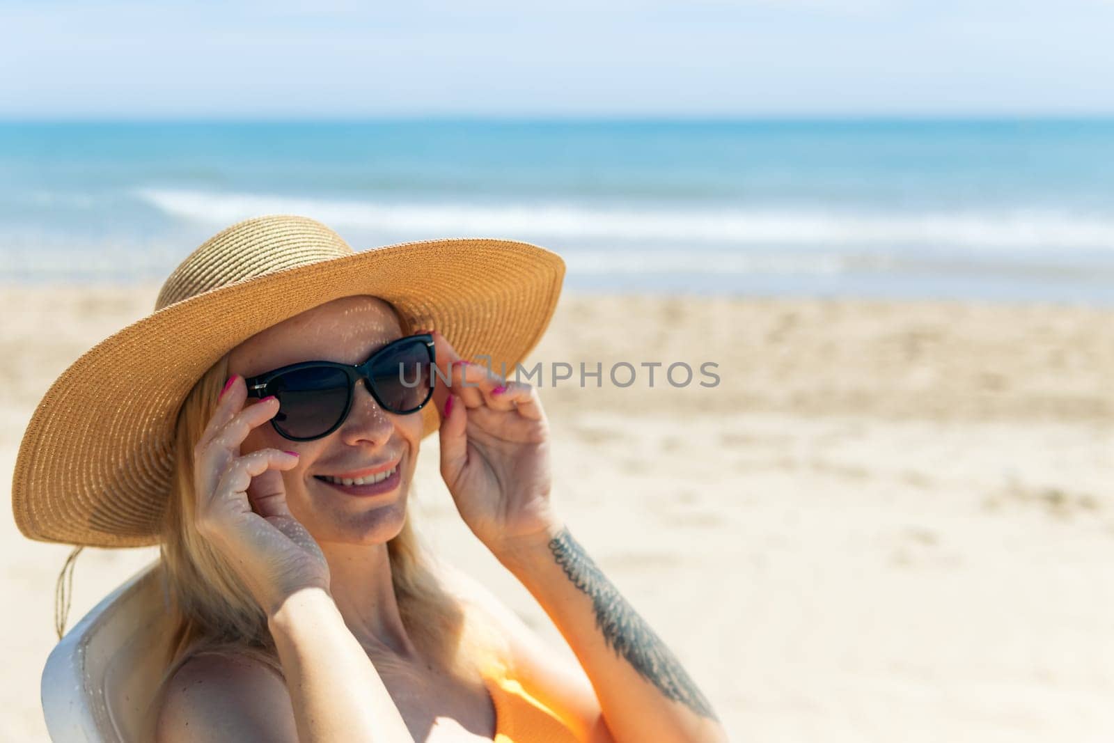 a beautiful seascape with a girl in a swimsuit and a hat, the girl smiles at the camera and holds sun glasses with her hands, the girl has a tattoo on her left arm. High quality photo