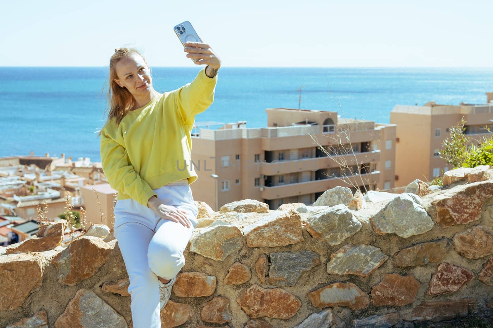 a girl with blond hair holds a phone in her hands and takes a selfie overlooking the sea, the girl is dressed in a yellow jacket and white pants by PopOff