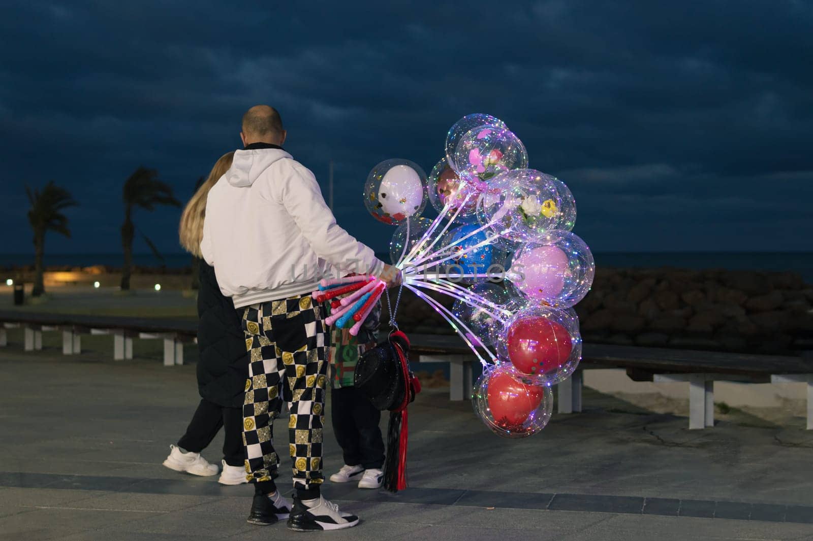 the guy stands with his back to the camera and holds in his hand a lot of multi-colored balloons of different colors and wants to give them to the girls on the waterfront by PopOff