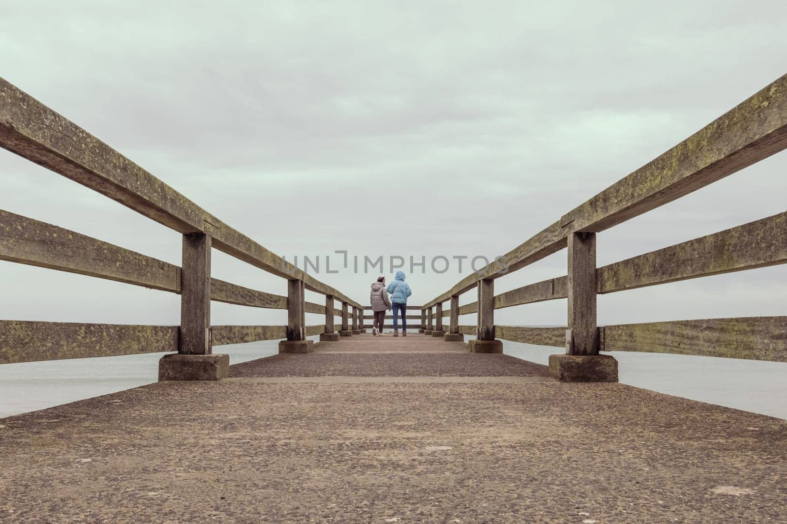 two girls stand in jackets on a bridge in the distance and look at the ocean, stand with their backs to the camera, there is a place for an inscription. High quality photo