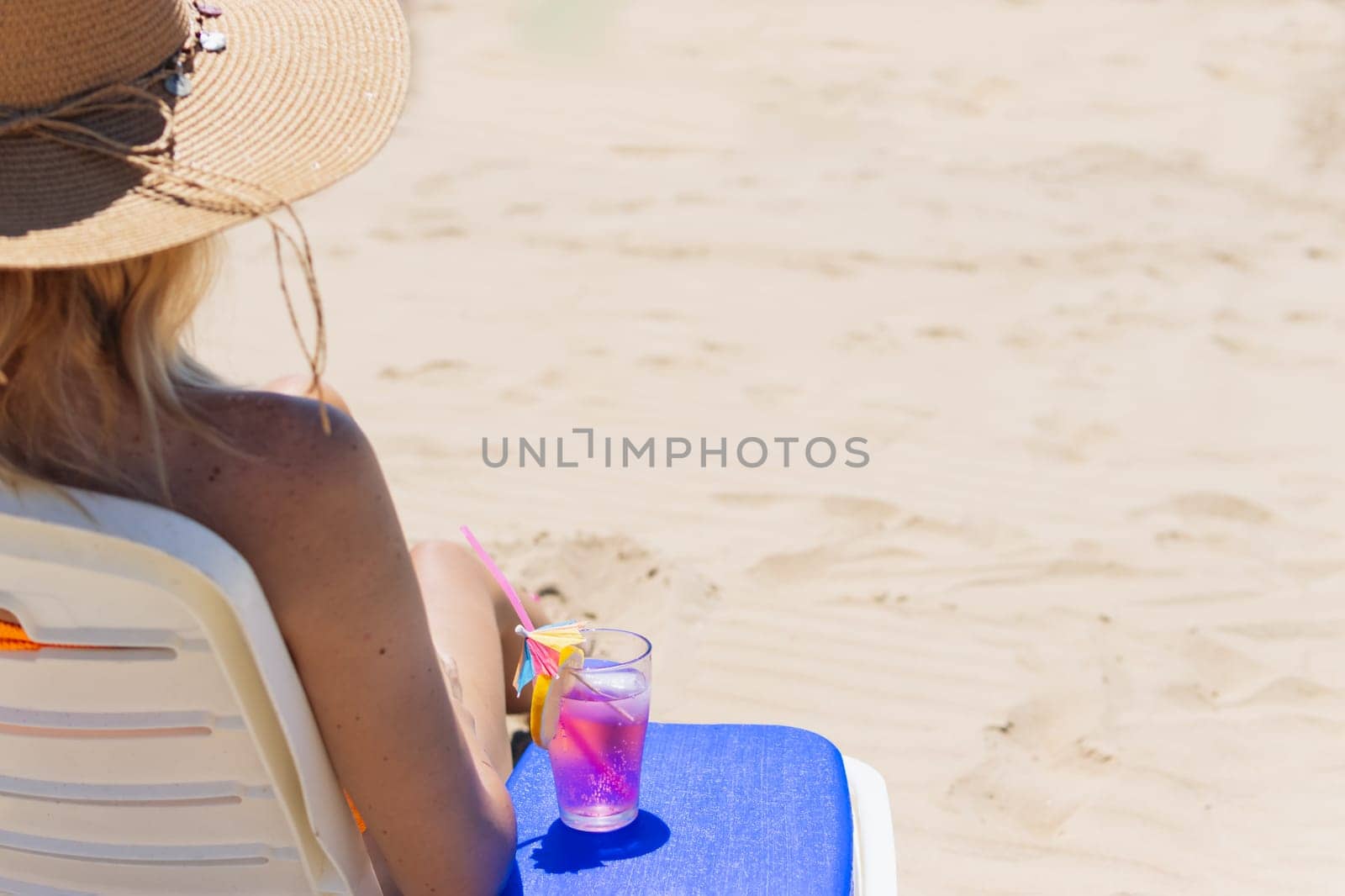 A sexy blonde in a bright bikini is sunbathing on a sun lounger with a cocktail in her hand by the sea. A beautiful girl is enjoying her summer holidays at an expensive resort, High quality photo