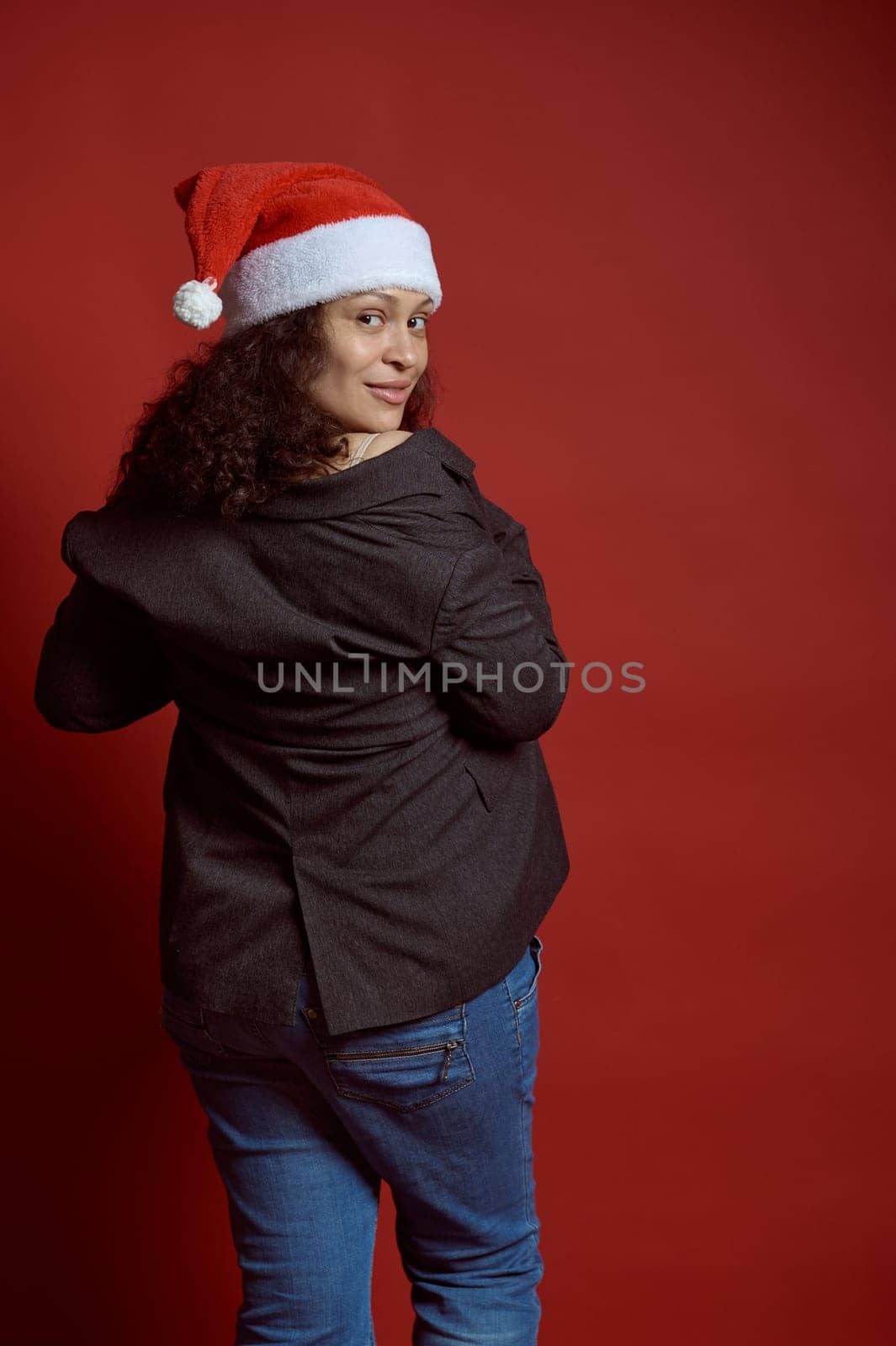 Back view of a beautiful multi ethnic young adult pregnant woman, wearing Santa hat, blue jeans and gray blazer, smiles cutely looking at camera, isolated over red color studio background