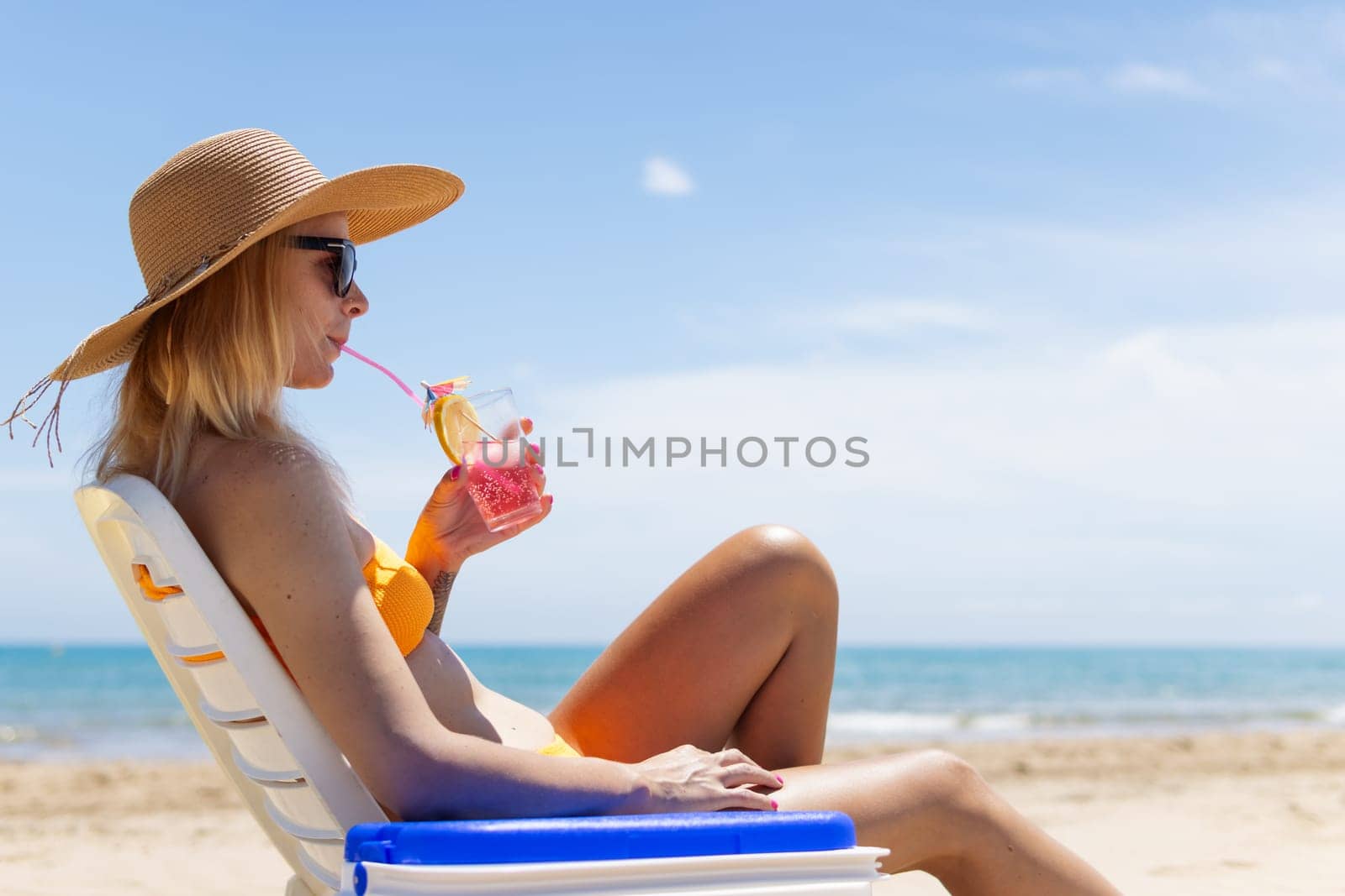 Happy young european girl with a cocktail in her hands is resting in a sun lounger on the sea near the blue refrigerator, there is space for an inscription. High quality photo