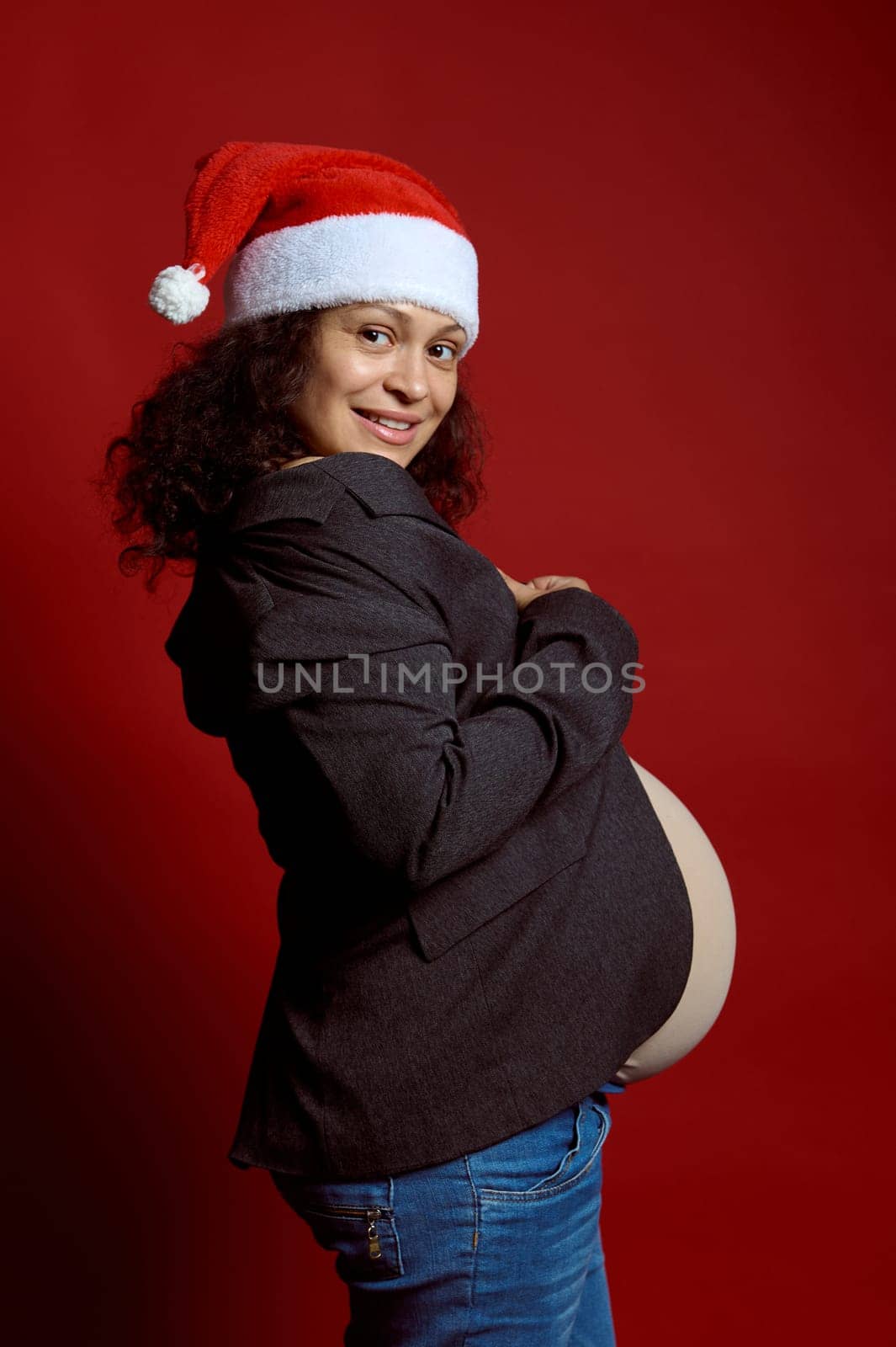 Authentic pregnant woman in Santa hat, smiles at camera, expressing positive emotions, isolated red studio background by artgf