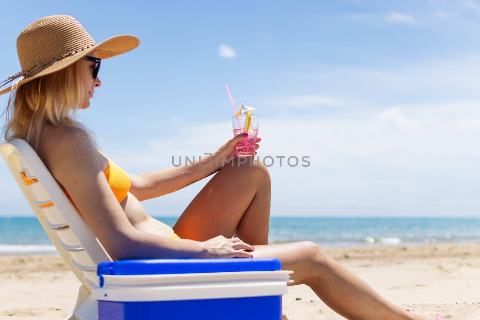 a slender blonde in a bright bikini, sunglasses and a hat is sunbathing on a sun lounger with a pink cocktail in her hand by the sea, next to a portable drinks fridge. . High quality photo