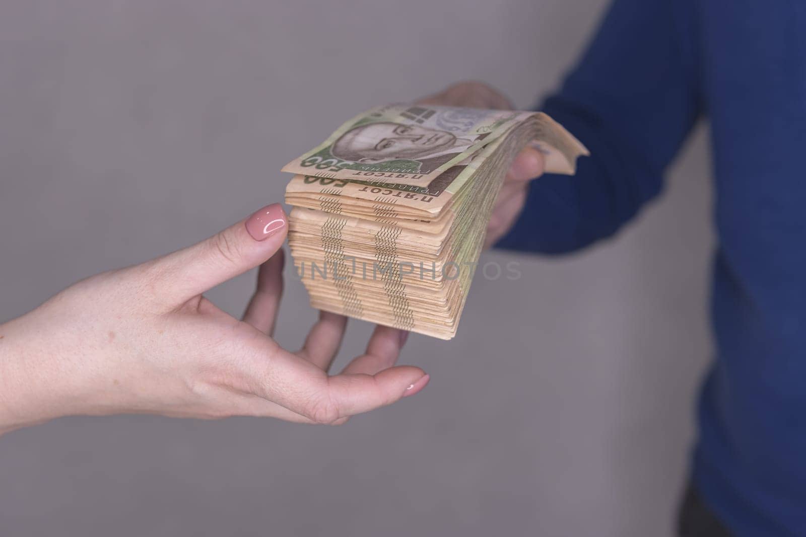 hands giving money isolated on a gray background.Finance concept.A man in a blue waiter gives a stack of money of 500 hryvnia to a woman's hands on a gray background. High quality photo