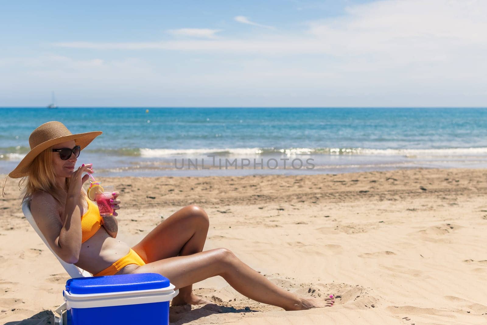 a slender blonde in a bright bikini, sunglasses and a hat is sunbathing on a sun lounger with a pink cocktail in her hand by the sea, next to a portable drinks fridge. . High quality photo