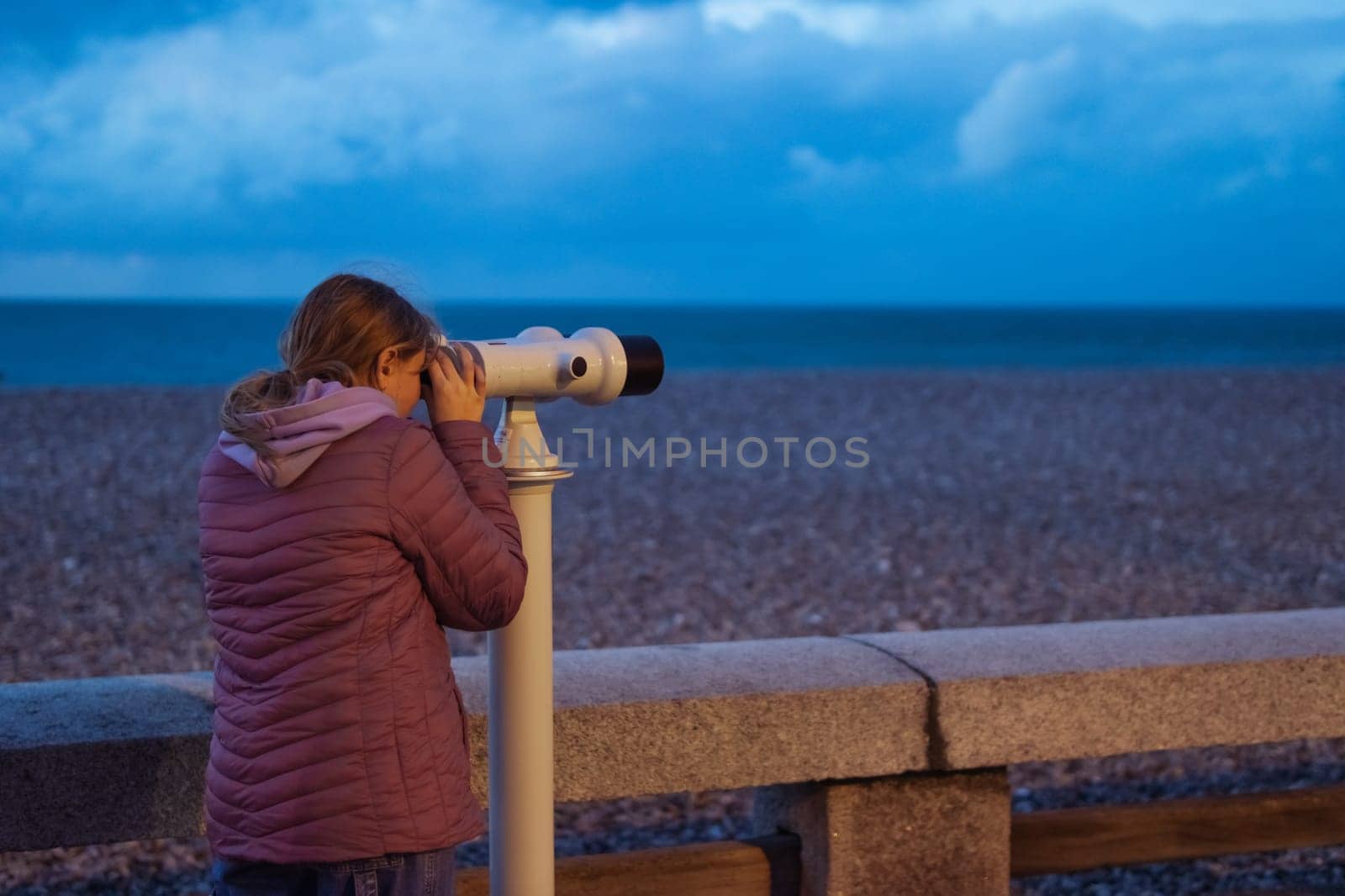 a girl of European appearance with blond hair stands on the embankment looking through binoculars at the ocean. the girl is dressed in a pink jacket and jeans, there is a place for. High quality photo