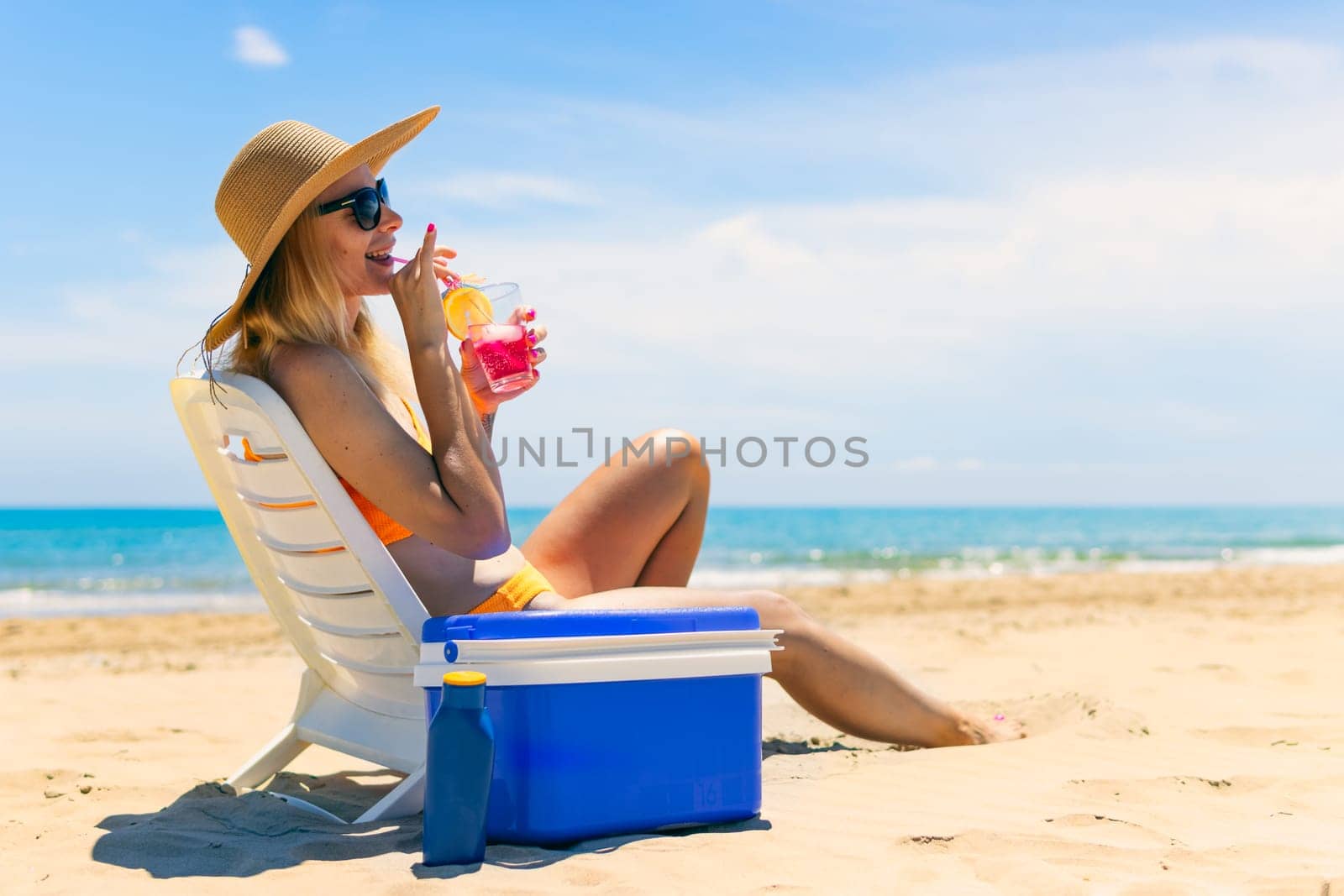 girl in a bathing suit, a hat sits on a sun lounger with a cocktail side view on the beach and there is a blue portable refrigerator for drinks next to it. Summer vacation concept. High quality photo