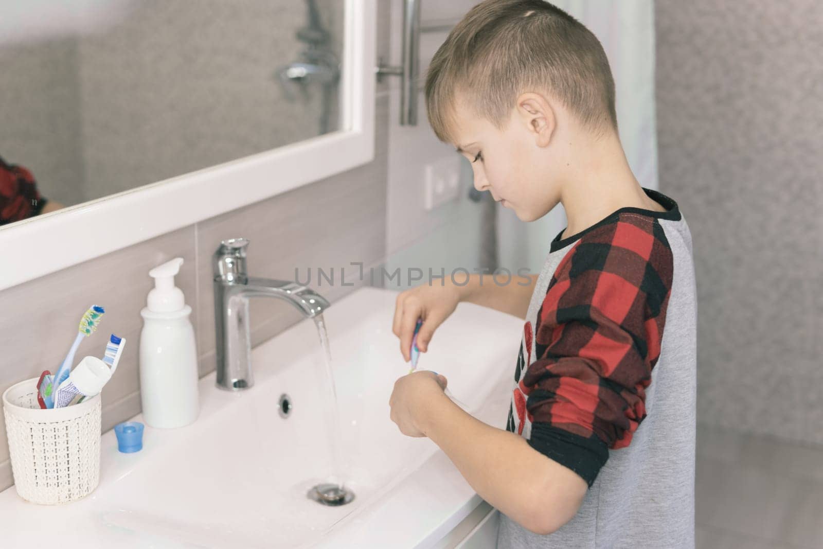 A small blond boy of European appearance learns to brush his teeth in a home bath in red pajamas and looks into a white mirror. Takes toothpaste. The child learns to stay healthy. healthcare concept.