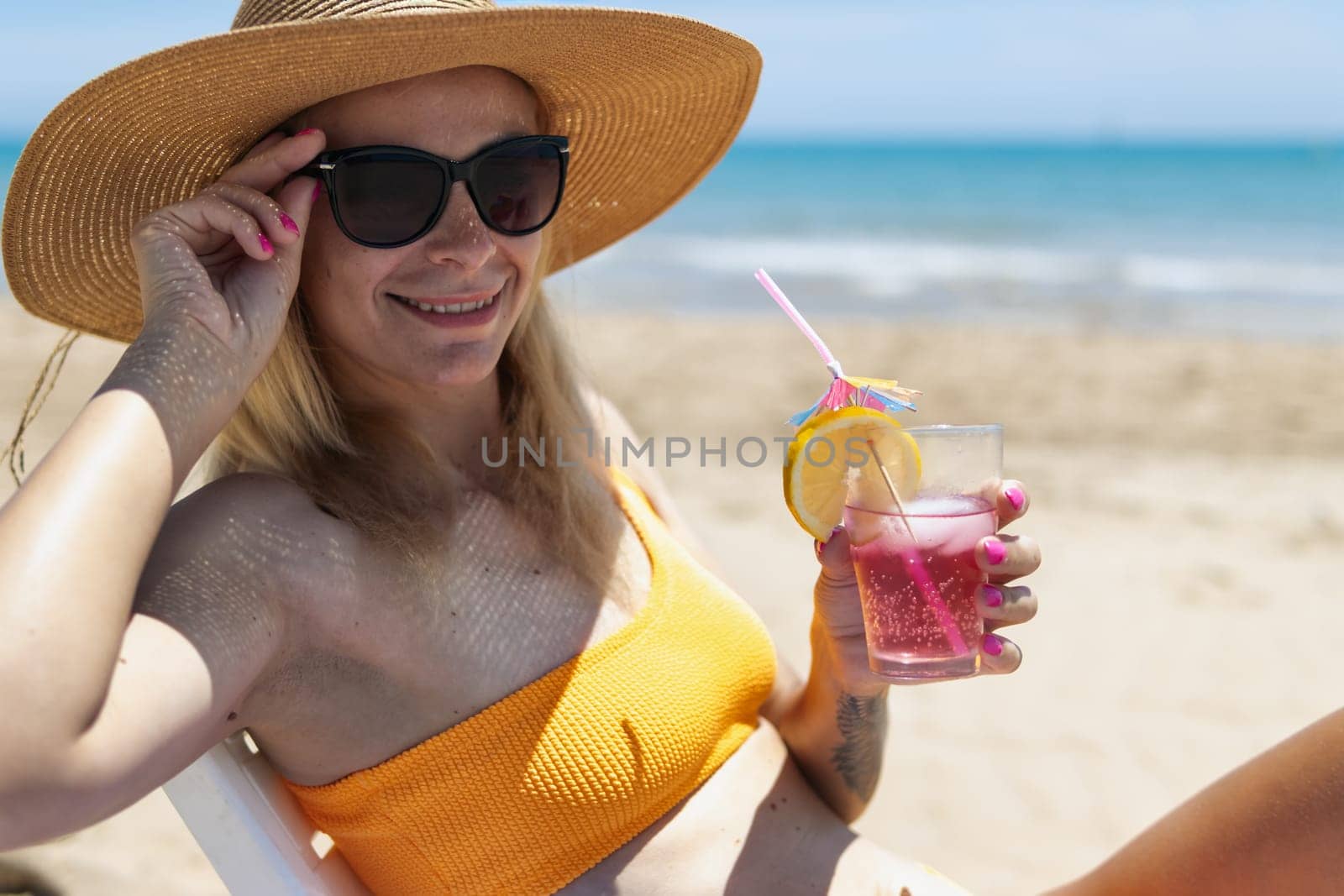 slender blonde in a bright bikini,sunglasses and is sunbathing on a sun lounger with a pink cocktail by PopOff