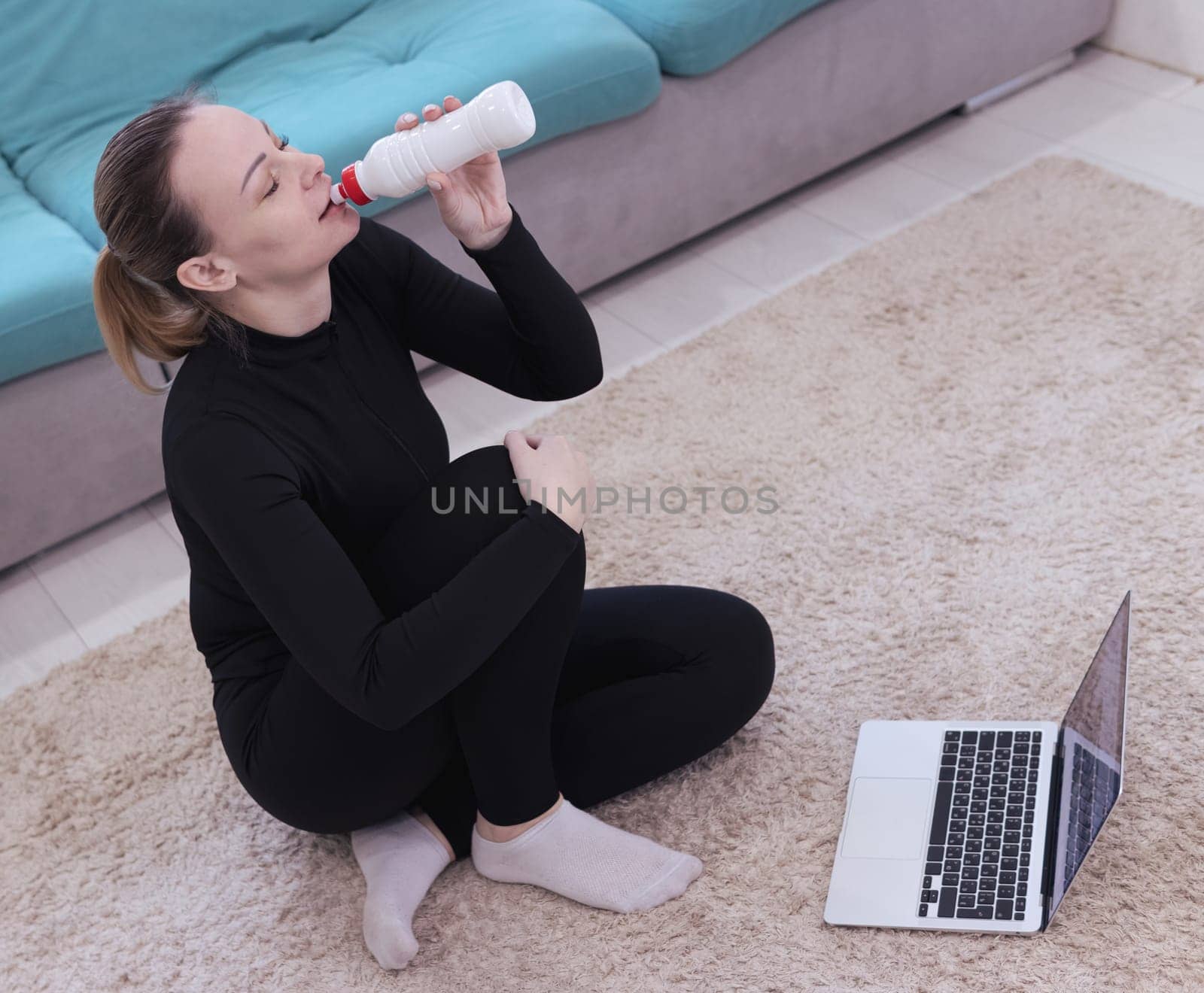A middle-aged athletic girl with blond hair with a ponytail, Caucasian nationality in black sportswear sits on the floor with dumbbells and drinks water from a bottle uses a laptop at home.