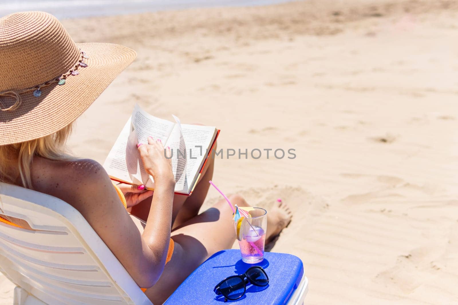 a slender girl in a swimsuit sits on a sun lounger sunbathes and reads a book on the beach, next to it is a blue portable refrigerator for drinks on it, a cocktail and glasses lie. High quality photo