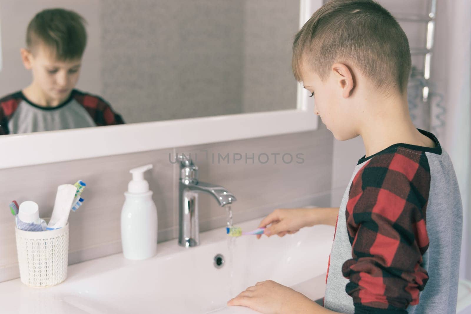 A small blond boy of European appearance learns to brush his teeth in a home bath in red pajamas and looks into a white mirror. The child learns to stay healthy. healthcare concept.