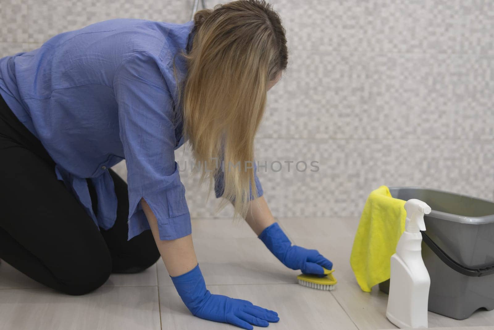 a blond girl with a European appearance in blue gloves cleans the tiles in the bathroom. in the hands of a yellow brush and next to a bucket with water and cleaning products.The concept of cleaning