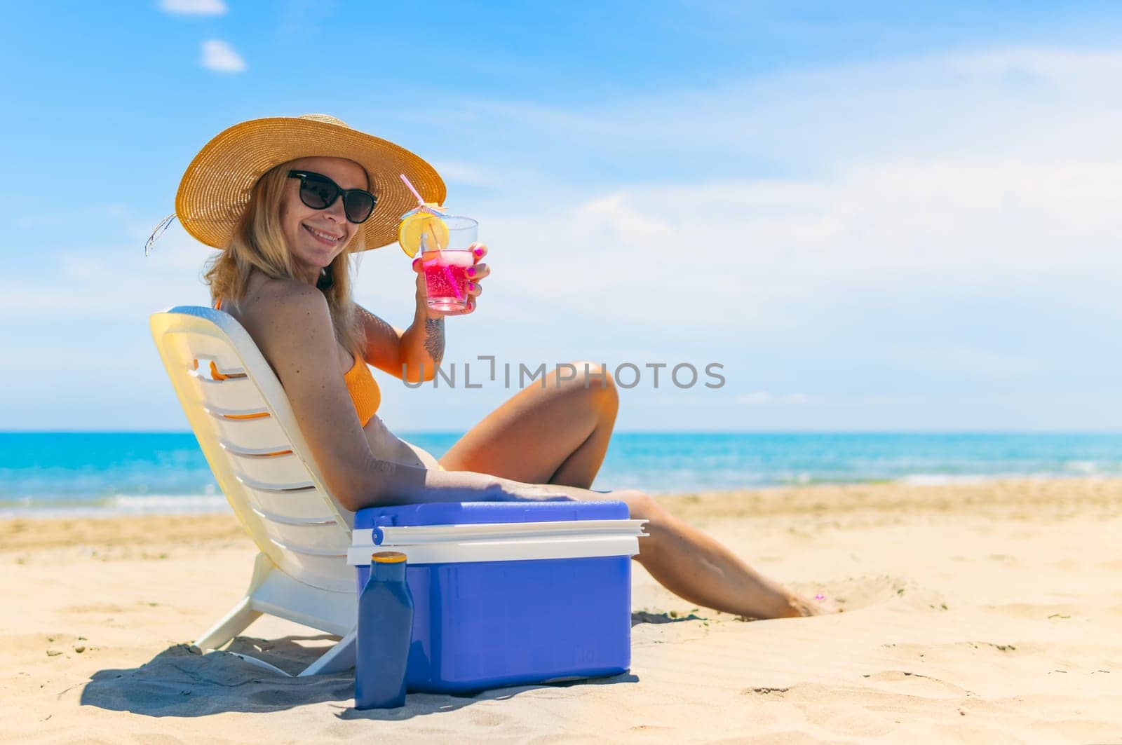 a cheerful girl in a bathing suit, a hat sits on a sun lounger with a cocktail in her hands side view on the beach and there is a blue portable refrigerator for drinks next to it. High quality photo