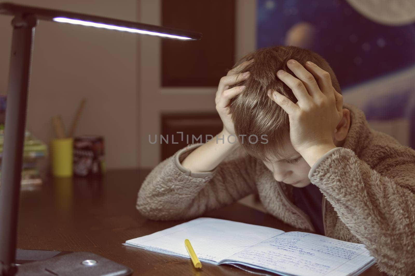 a boy with a European appearance with blond hair is focused on doing homework in a children's room on distance learning in front of the phone and writing in a notebook with a pen