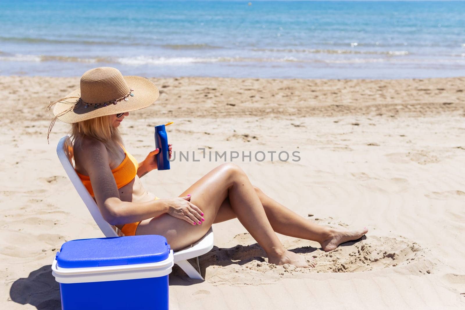 a tanned girl in a swimsuit sits on a sun lounger smears sunblock on her legs side view on the beach by PopOff