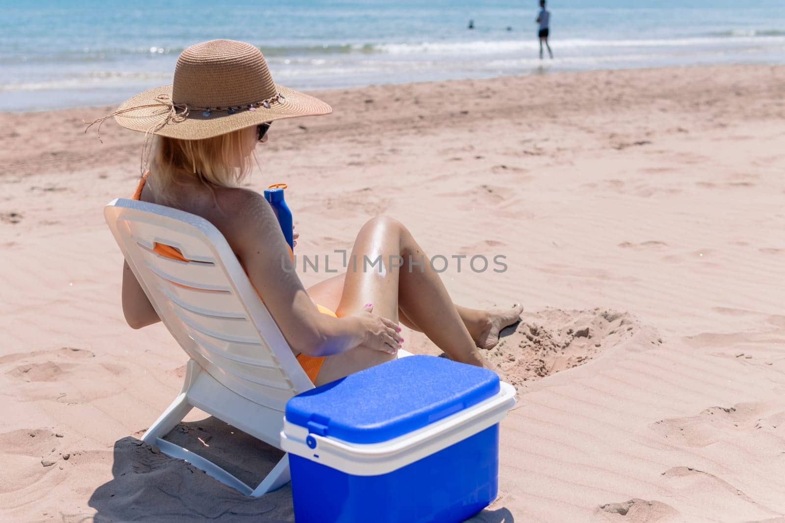 a tanned girl in a swimsuit sits on a sun lounger smears sunblock on her legs rear view on the beach and a blue portable refrigerator for drinks stands nearby.Summer vacation High quality photo