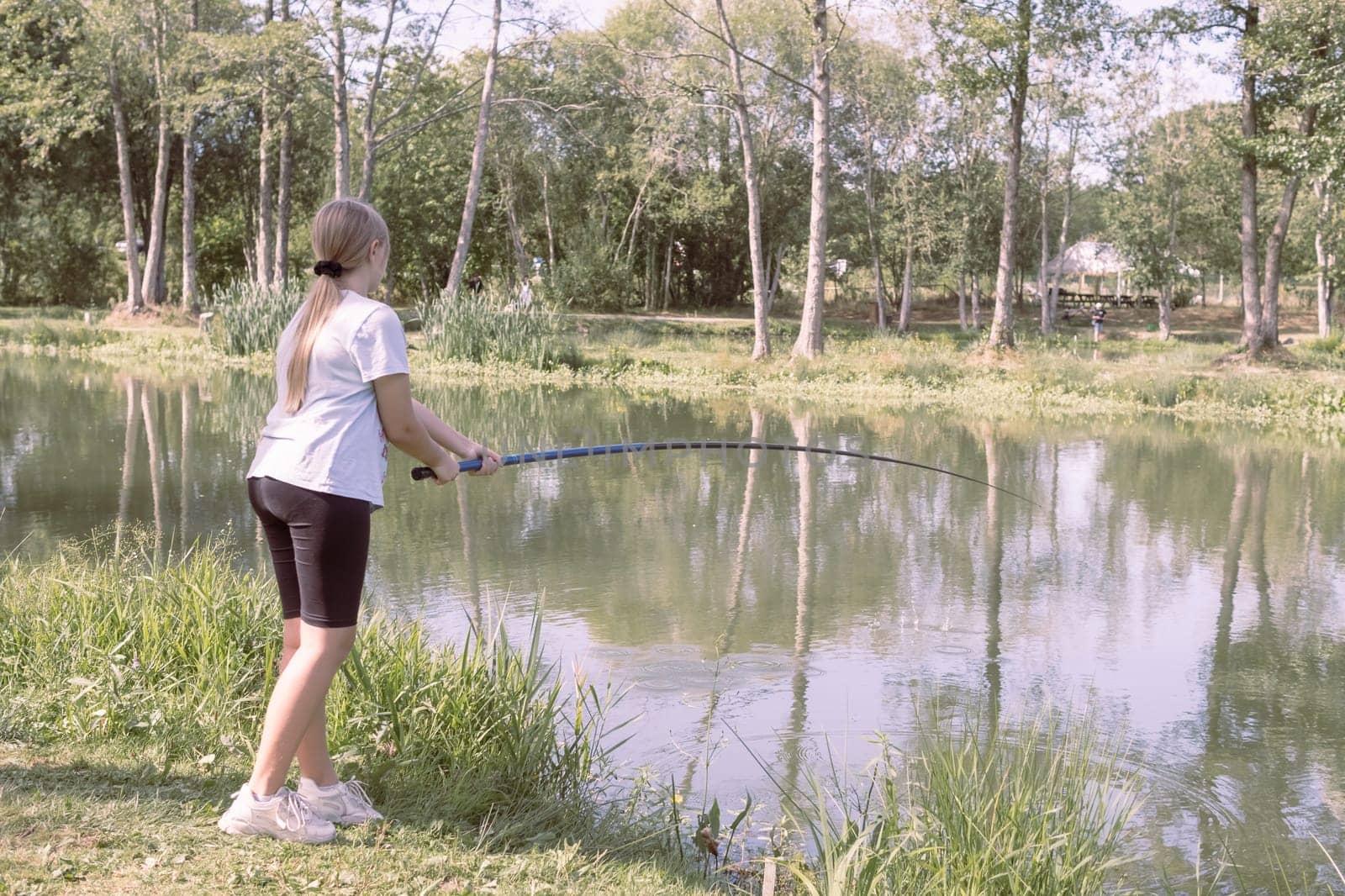 a teenager girl of European appearance with blond hair tied in a ponytail, stands fishing at a stake by PopOff