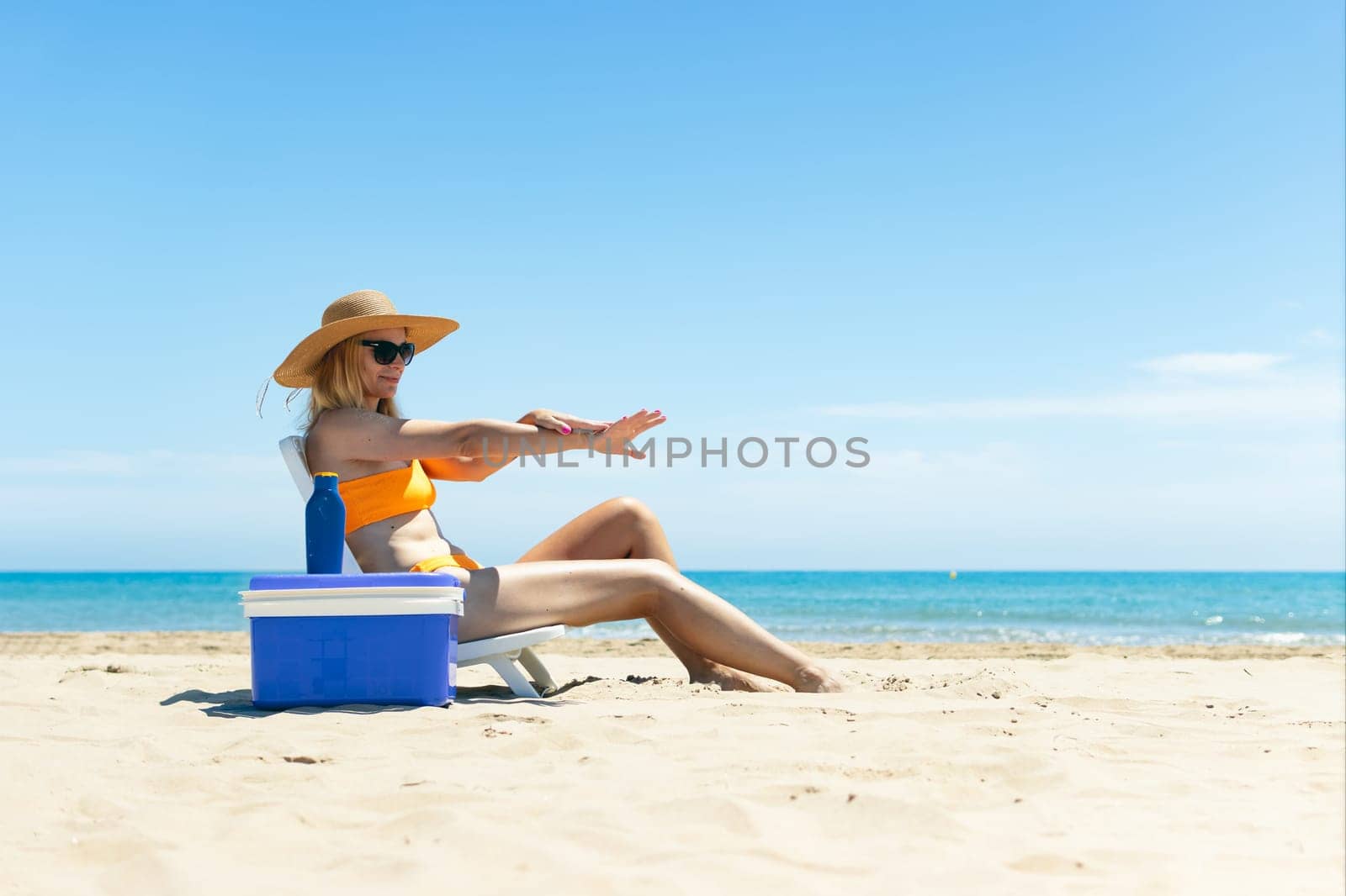 a tanned girl in a swimsuit, hat and glasses sits on a sun lounger smears sunscreen on her hands by PopOff
