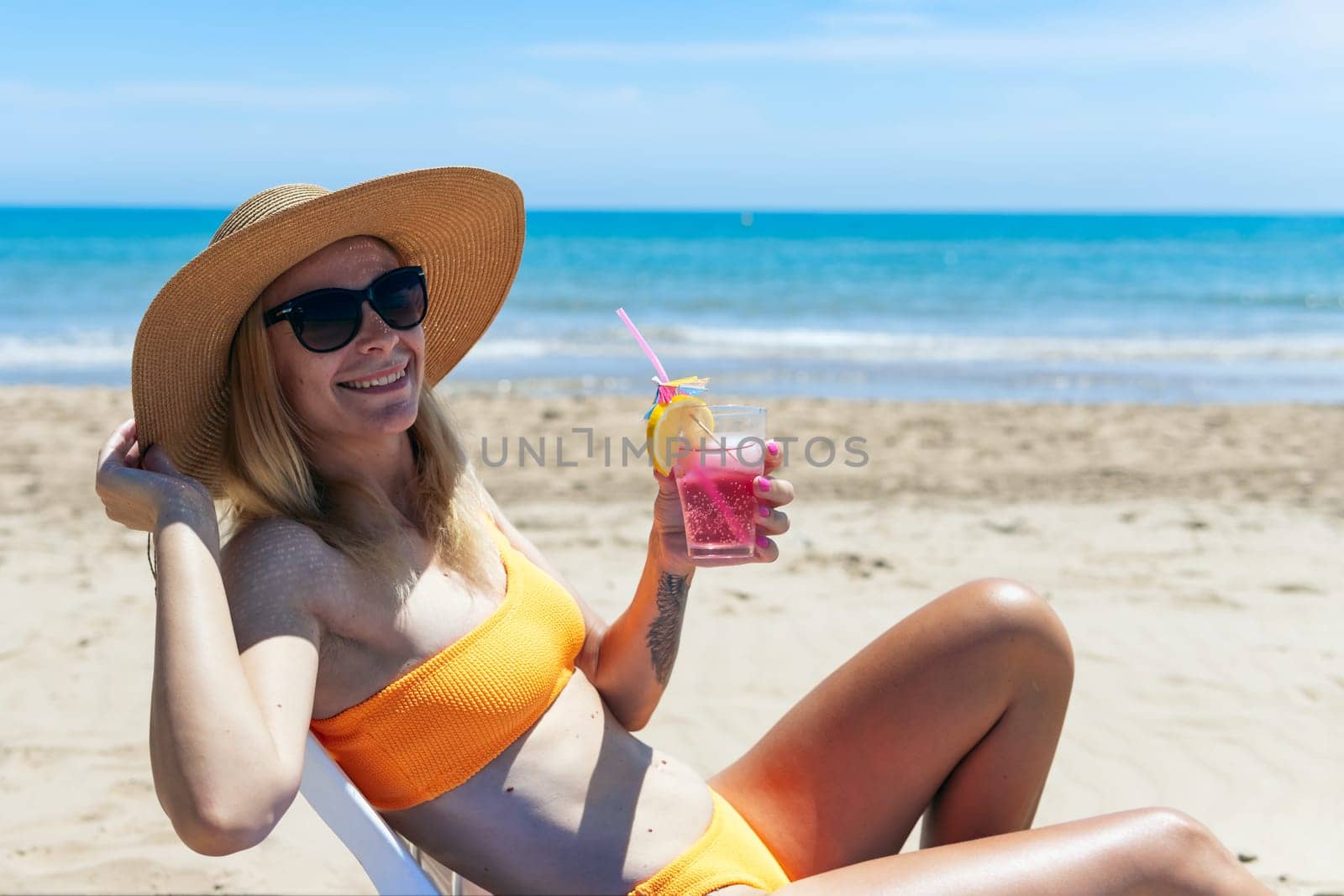 A sexy blonde in a bright bikini is sunbathing on a sun lounger with a cocktail in her hand by the sea. A beautiful girl is enjoying her summer holidays at an expensive resort, High quality photo