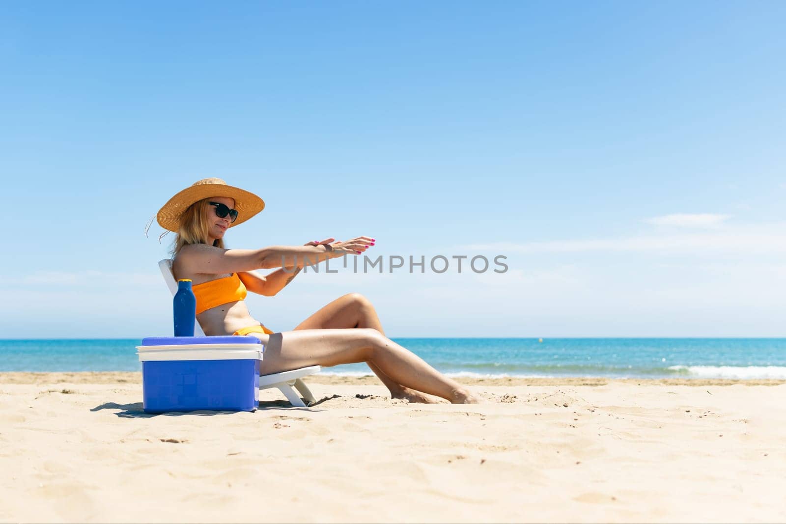 a tanned girl in a swimsuit, hat and glasses sits on a sun lounger smears sunscreen on her hands by PopOff