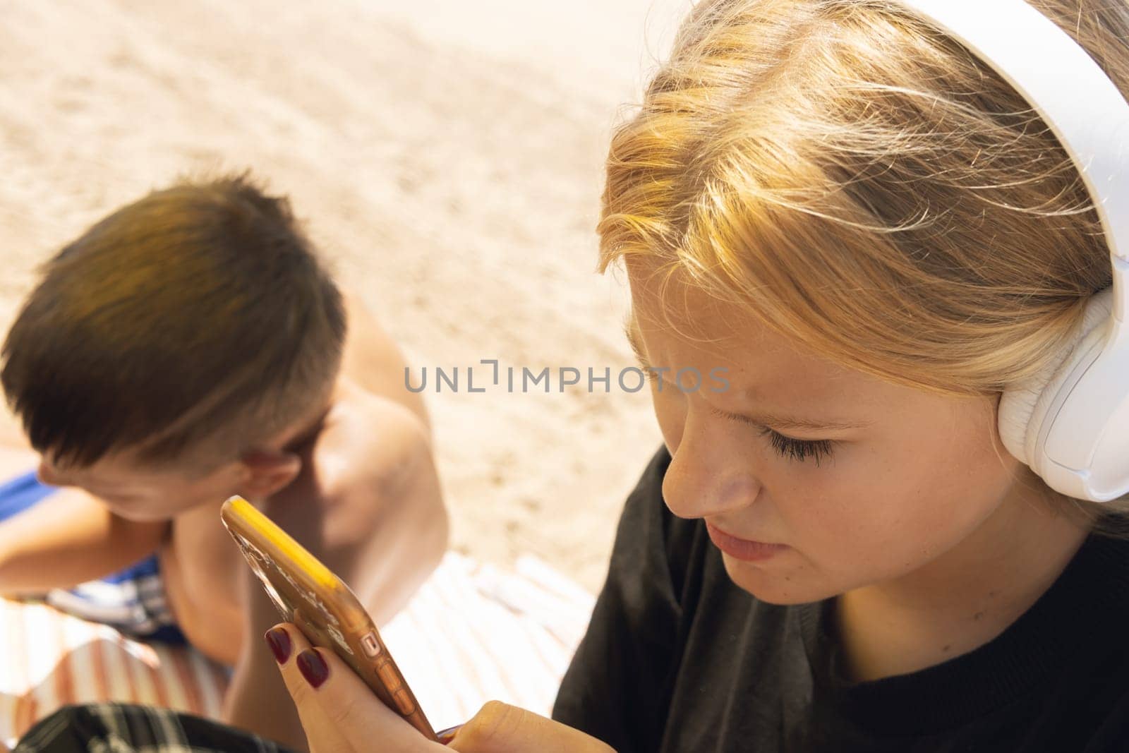 teenagers boy and girl are sitting on the beach, the girl is wearing headphones and with a phone in her hands listening to music, the boy is lying sunbathing. High quality photo