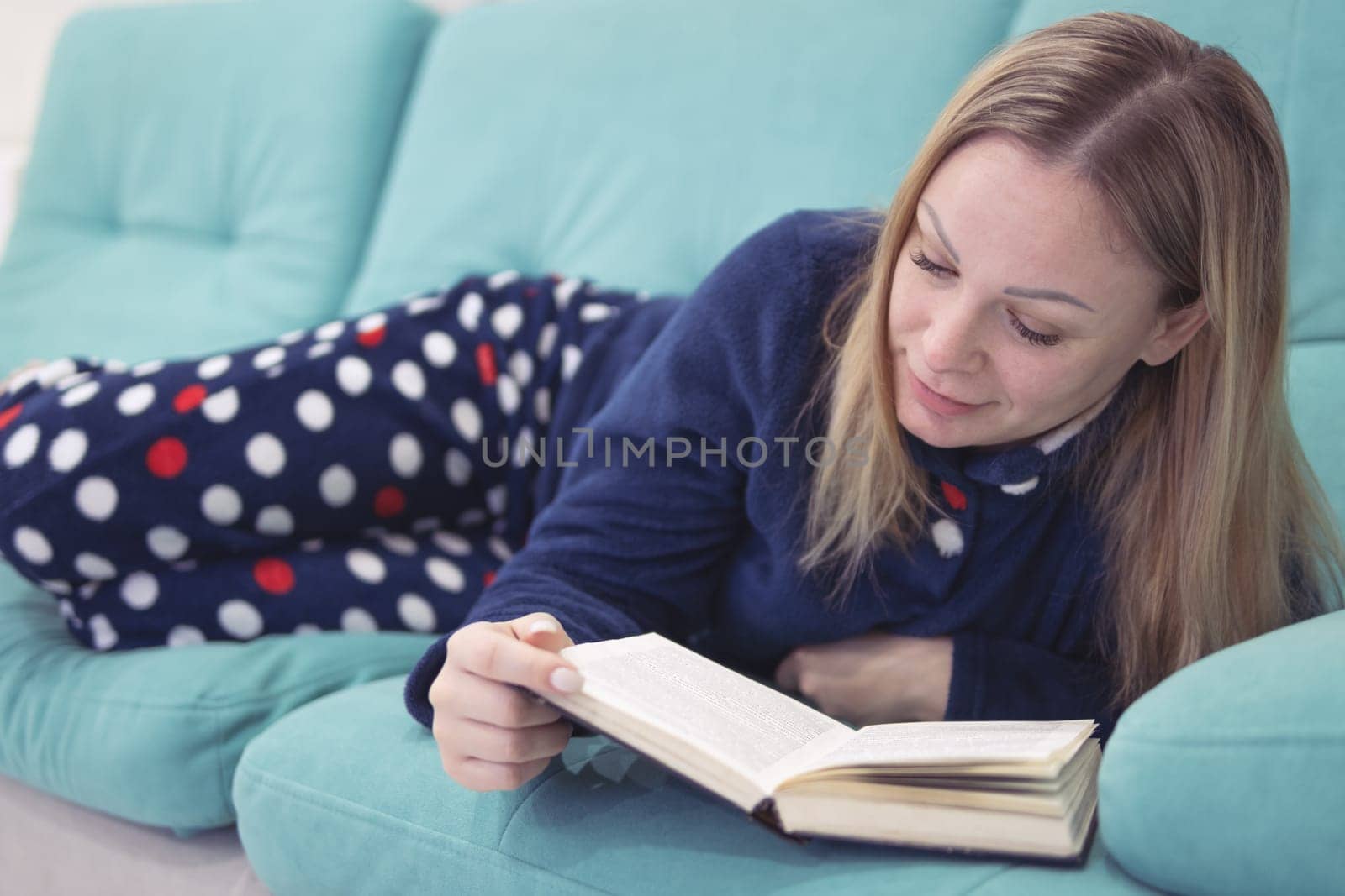 A girl with blond hair of European appearance smiling reads a book while lying on a green sofa in the living room in blue pajamas. The concept of relaxation and reading.