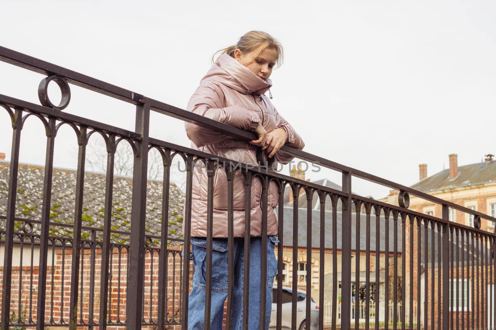 a teenage girl of European appearance with blond hair in a pink jacket and jeans stands on a bridge in the park close-up, the child looks to the side by PopOff