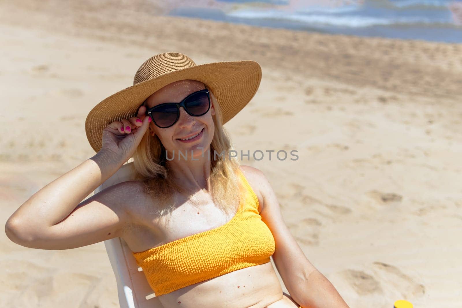 Portrait of a happy european woman in an orange swimsuit and a hat and glasses sunbathing on a plastic sun lounger on the beach.Girl with her hand raised up is sunbathing on the sea.High quality photo