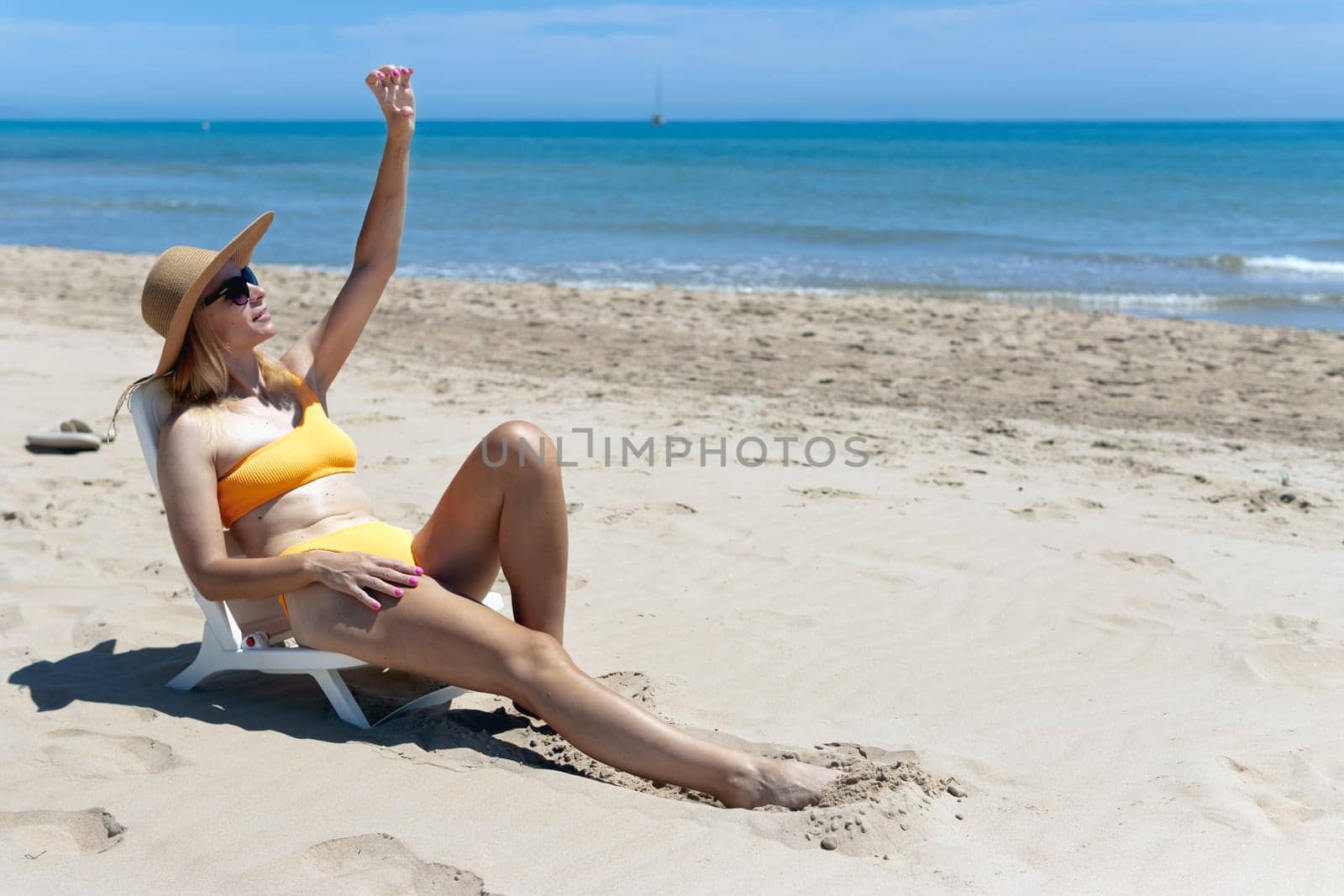 Portrait of a happy european woman in an orange swimsuit and a hat and glasses sunbathing on a plastic sun lounger on the beach.Girl with her hand raised up is sunbathing on the sea.High quality photo