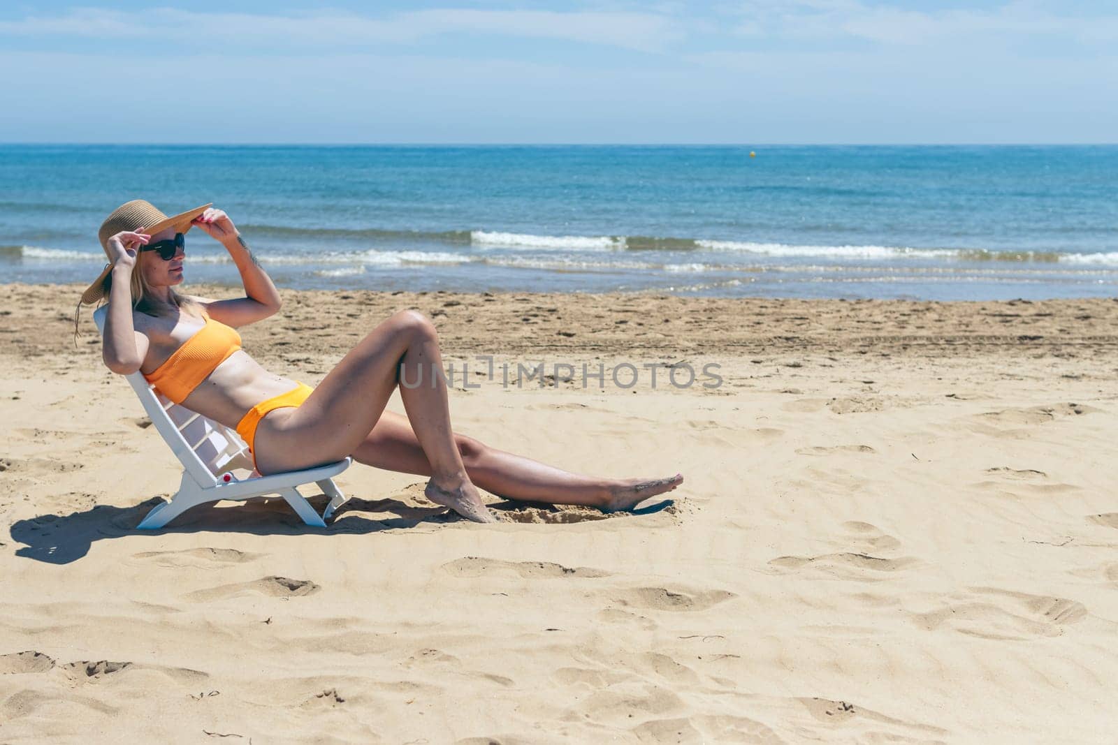 european girl in a hat and swimsuit relaxes and lies on a sun lounger on the beach alone, by PopOff