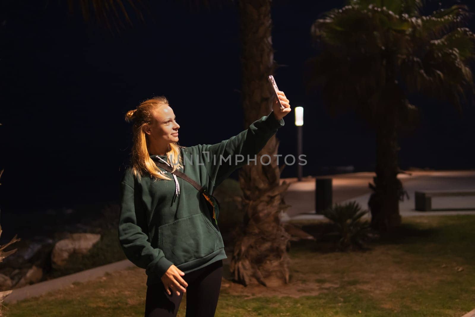 Close-up of a cheerful girl makes a selfie photo on a mobile phone on a night city street. Portrait woman showing victory sign to smartphone camera outdoors. high quality photo. High quality photo