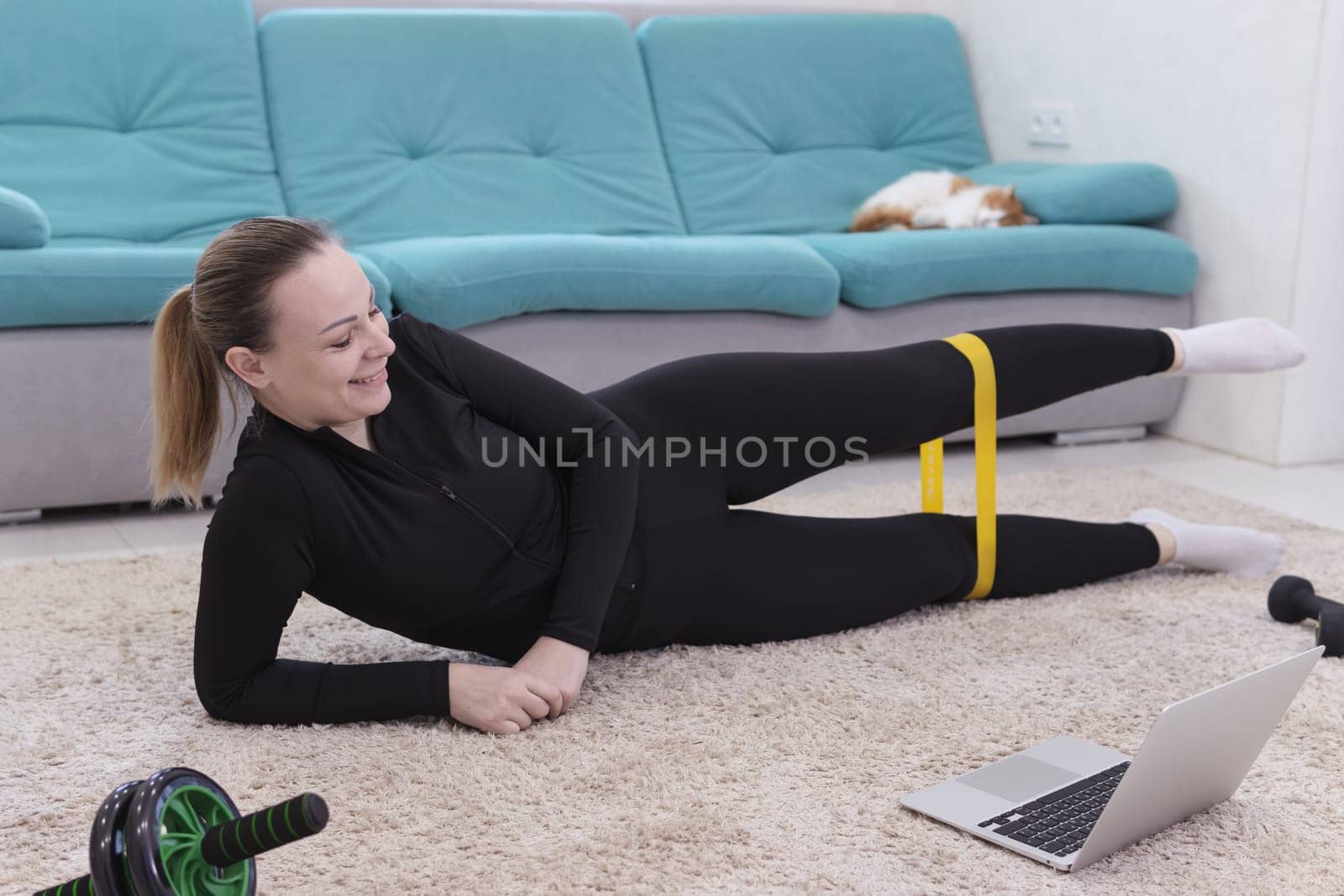 young smiling girl of European appearance with blond hair in a black sport uniform goes in for sport with a sport elastic band at home lying on the floor in a laptop.The concept of home training