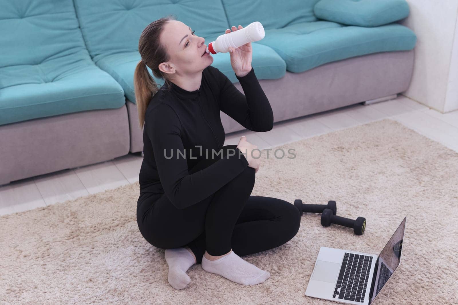 A middle-aged athletic girl with blond hair with a ponytail, Caucasian nationality in black sportswear sits on the floor with dumbbells and drinks water from a bottle uses a laptop at home.