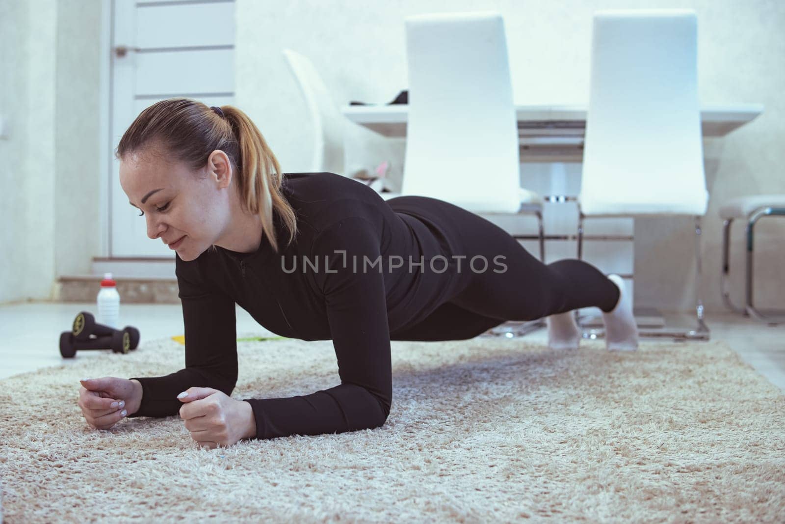 A beautiful young woman of European appearance in a sports uniform is doing a plank in the living room at home lying on a white carpet, in the background is a white dining table, chairs