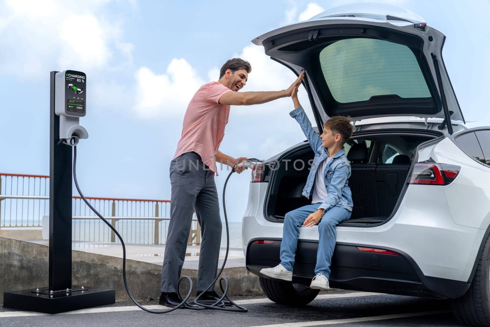 Family road trip vacation traveling by the sea with electric car, father and son high five after reach destination at EV charging station by the seashore. Eco-friendly car for environment. Perpetual