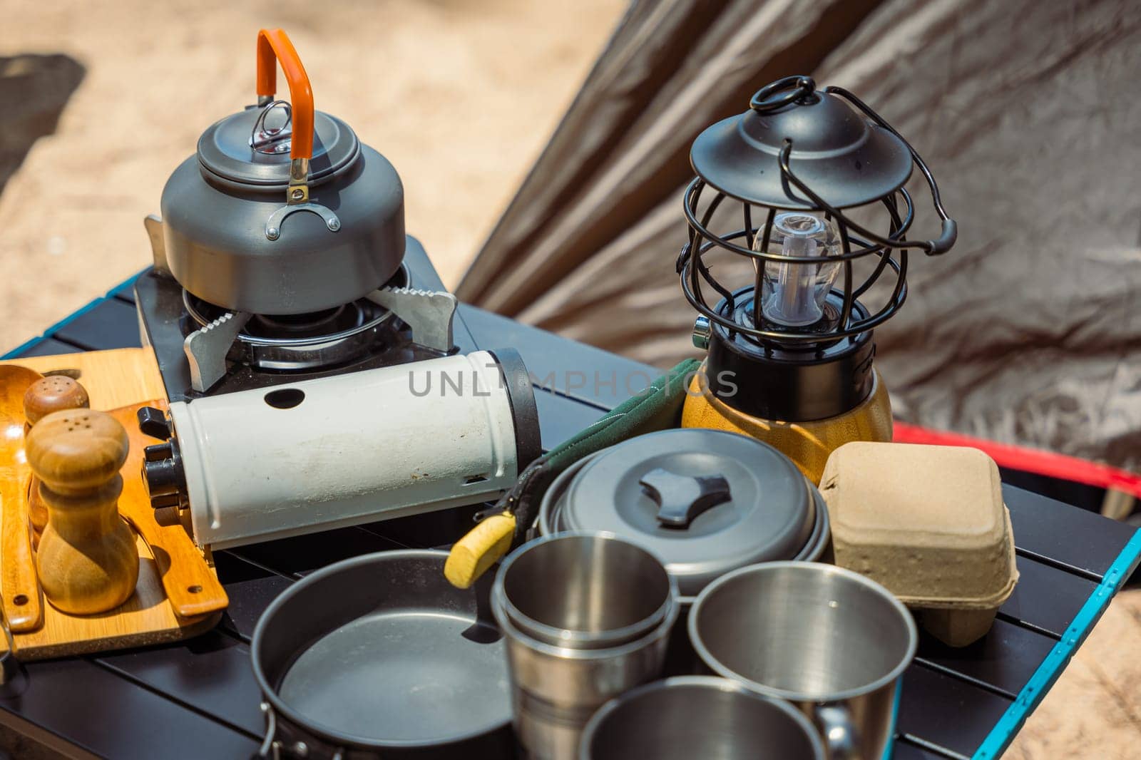 Camping essentials neatly arranged at a beachfront tent, kettle, pot, pan, gas stove, flashlight, and camera. Perfect setup for a relaxing outdoor journey in nature.