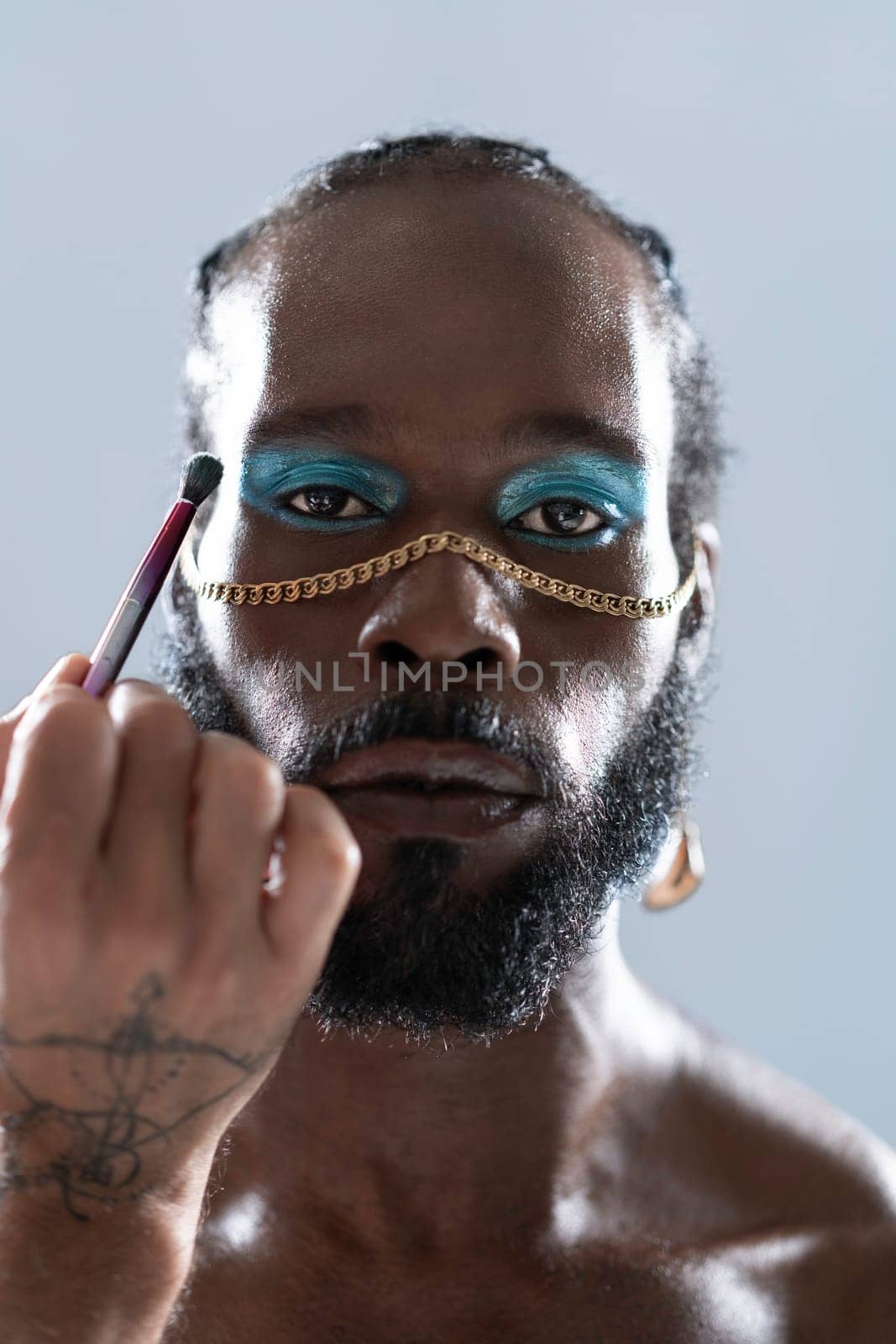 Confident black gay man with bright makeup and accessory. Standing isolated on light blue background. African-american homosexual man does brush makeup on his face. Cosmetics and gender identity