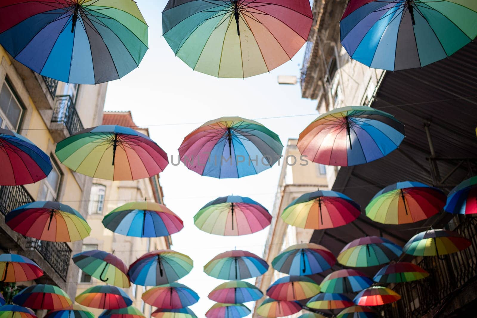 Colorful floating umbrellas hang above the street. Street decorated with colored parasols. Rainbow umbrellas on blue sky background. Many rainbow parasols, street decoration for festivals. Pride month