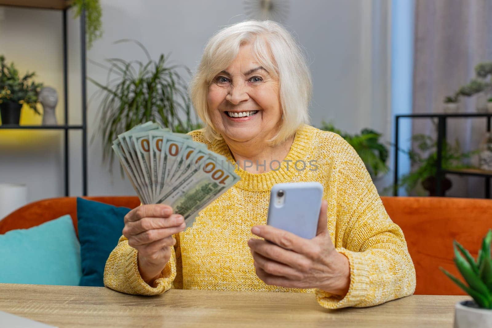 Planning family budget. Happy senior woman counting money cash use smartphone calculate domestic bills at home. Mature excited grandmother satisfied of income and saves money for planned vacation gift