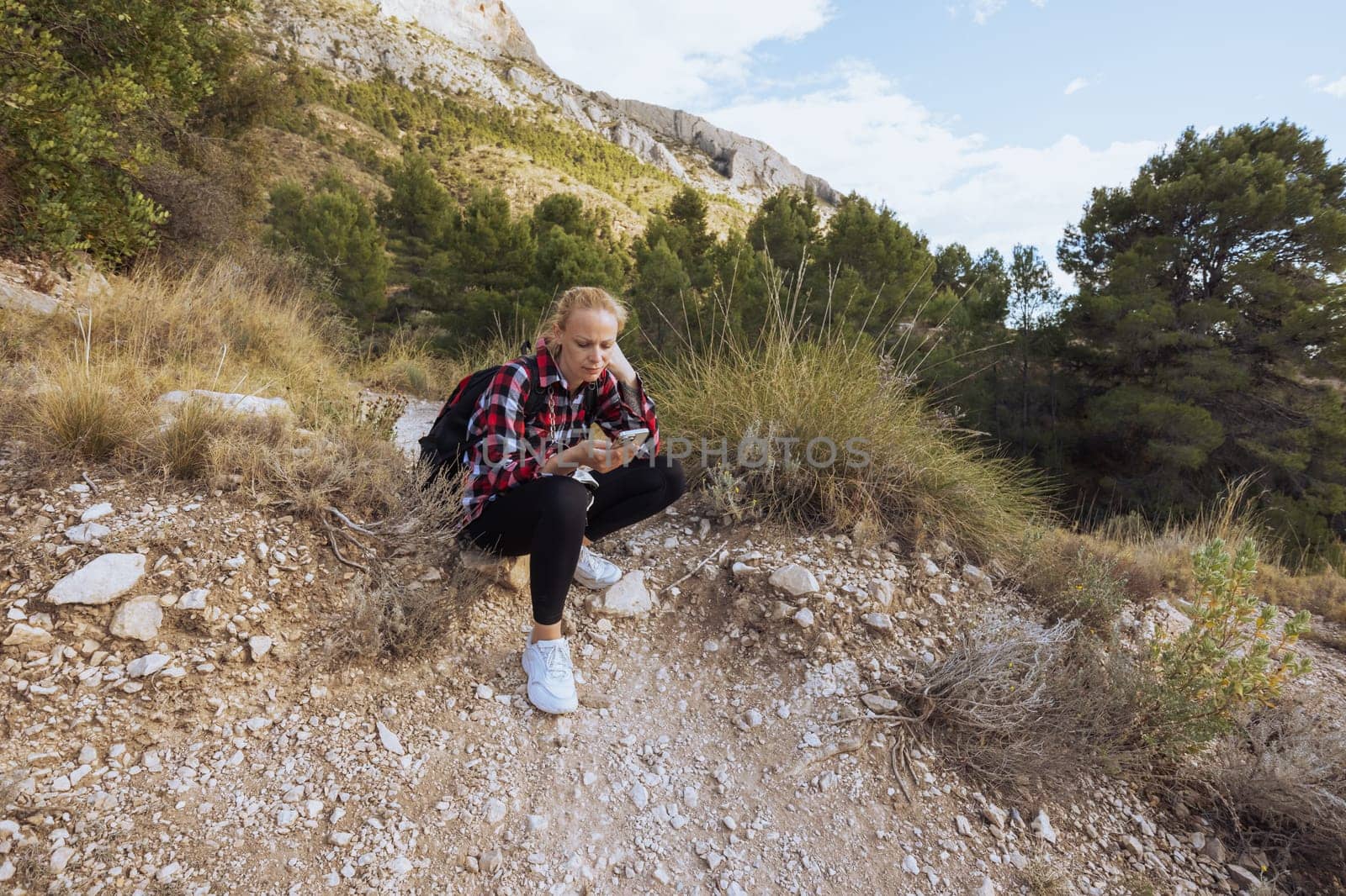 a girl in sportswear sits on a stone, rests from a hike in the mountains, a girl sits looking at the phone by PopOff