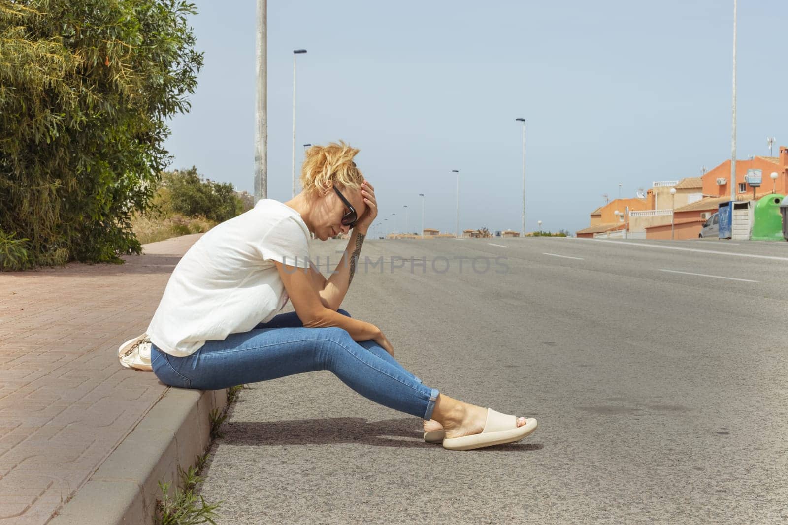 a sad girl sits on the sidewalk in jeans and a T-shirt and glasses, looks at the road by PopOff